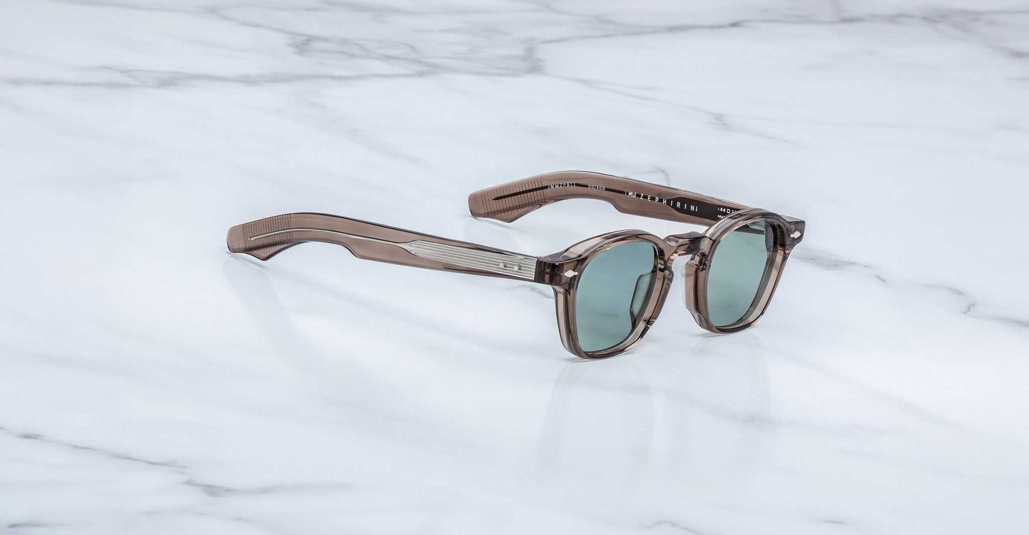 A pair of Jacques Marie Mage Zephirin sunglasses with brown translucent handcrafted acetate frames and round green lenses rests on a white marble surface.