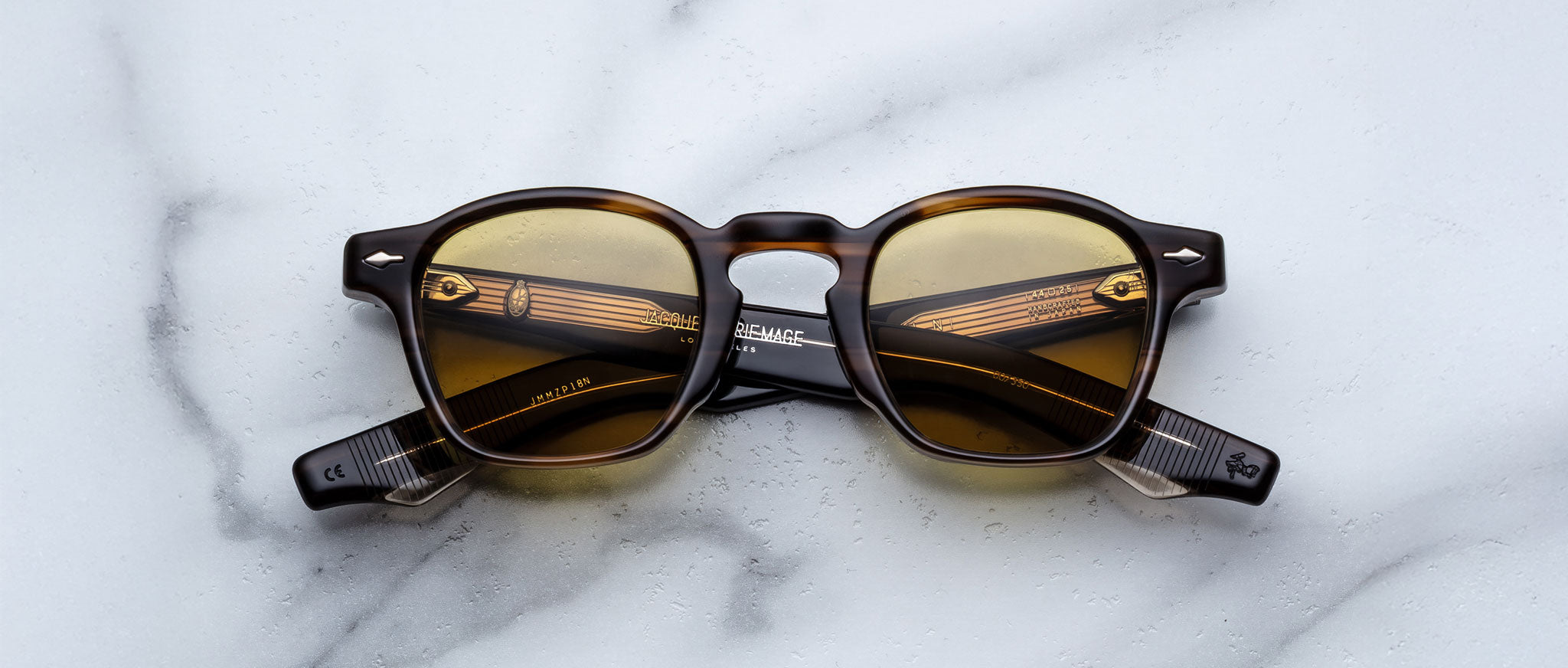 Jacques Marie Mage Zephirin round sunglasses with brown-tinted lenses and dark patterned frames are displayed on a white marble surface, highlighting the timeless elegance of this limited edition eyewear by Jacques Marie Mage.