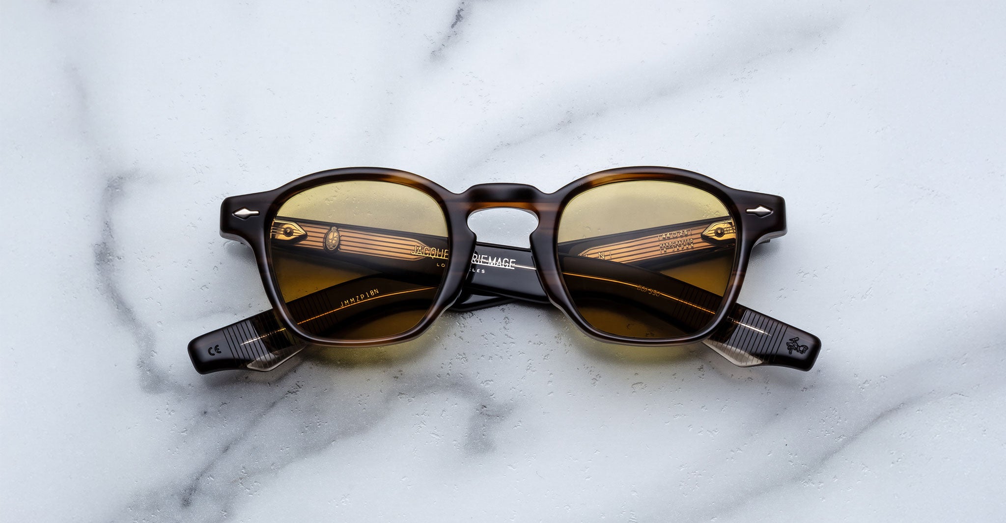 Jacques Marie Mage Zephirin round sunglasses with brown-tinted lenses and dark patterned frames are displayed on a white marble surface, highlighting the timeless elegance of this limited edition eyewear by Jacques Marie Mage.