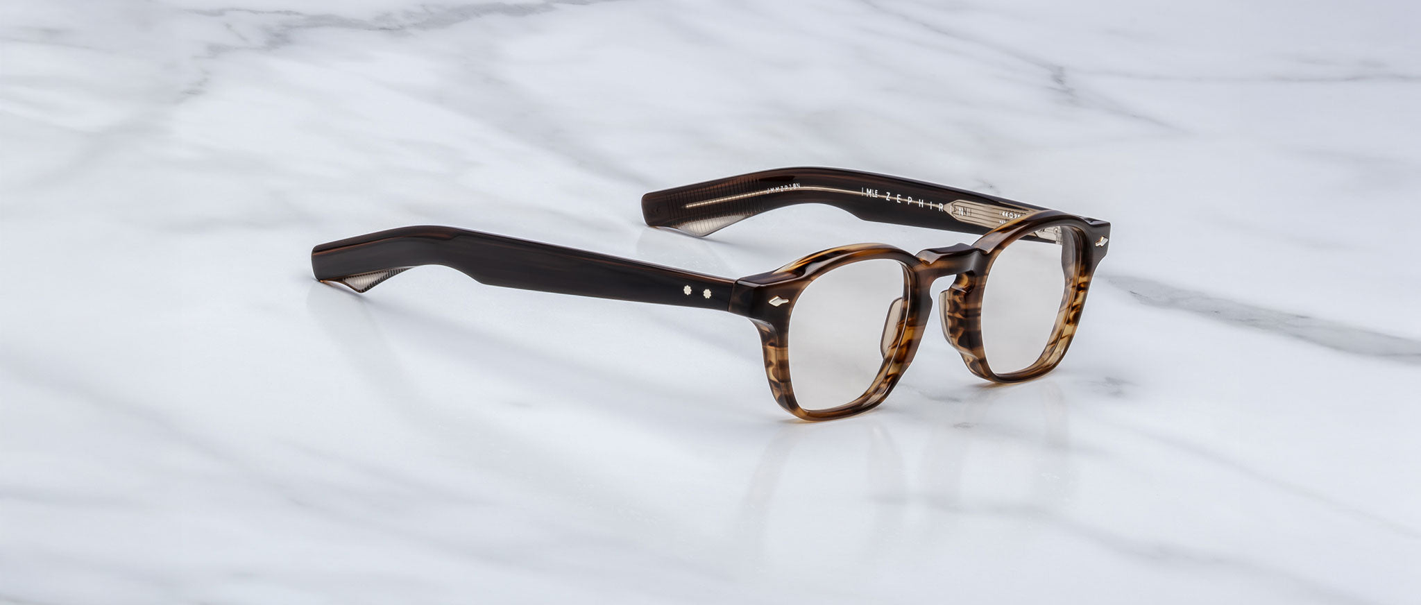 The Jacques Marie Mage Zephirin spectacles by Jacques Marie Mage, featuring handcrafted thick tortoiseshell acetate frames and clear lenses, are elegantly displayed on a white marble surface.