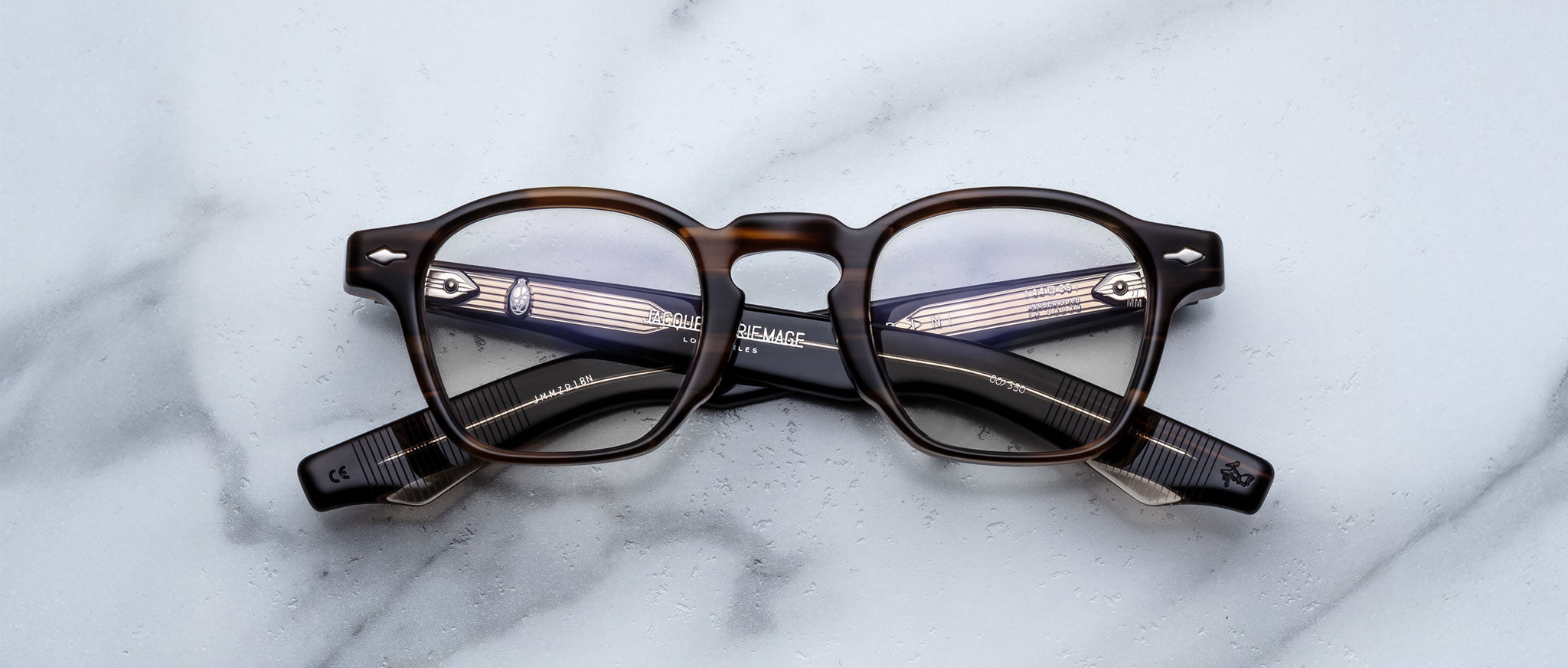 A pair of Jacques Marie Mage Zephirin spectacles with round lenses, handcrafted in acetate, is displayed on a white marble surface.