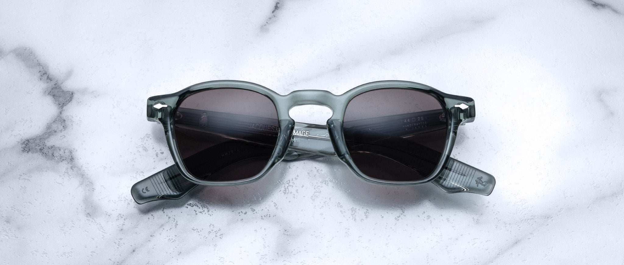A pair of Jacques Marie Mage Zephirin sunglasses with dark lenses and transparent gray frames rests on white marble, highlighting the sophistication of this limited edition Jacques Marie Mage eyewear.