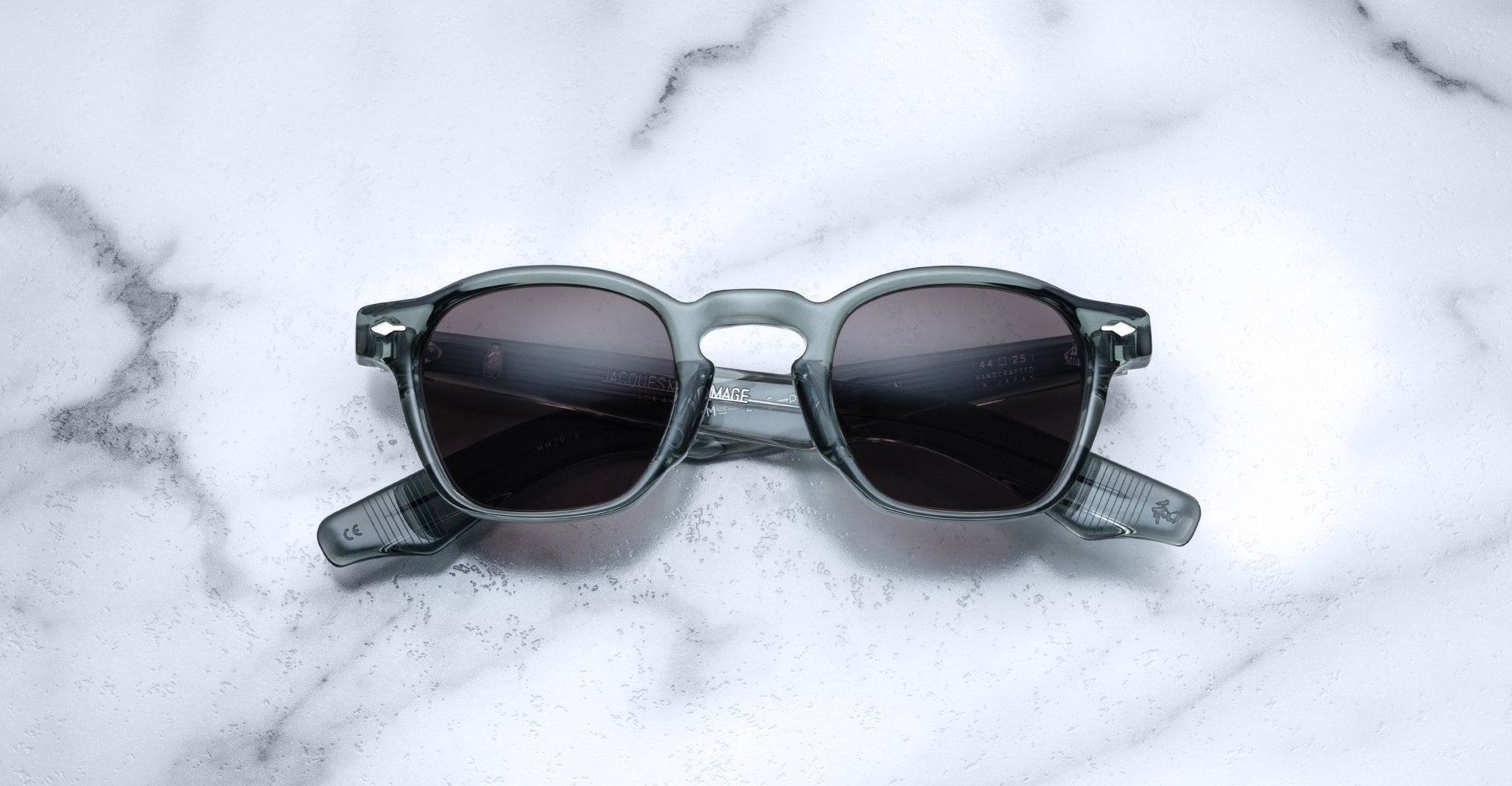A pair of Jacques Marie Mage Zephirin sunglasses with dark lenses and transparent gray frames rests on white marble, highlighting the sophistication of this limited edition Jacques Marie Mage eyewear.
