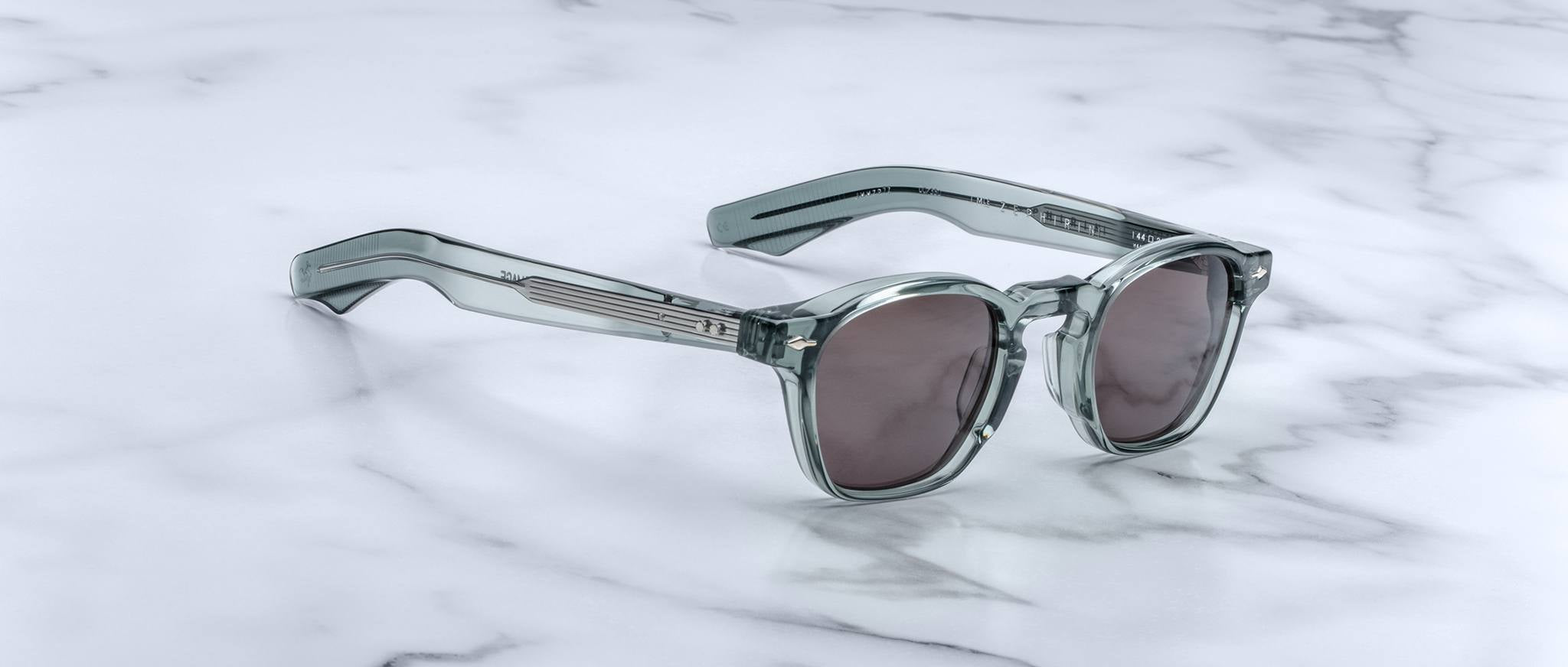 Gray sunglasses with thick translucent frames and dark lenses rest on white marble, evoking the limited edition Jacques Marie Mage Zephirin by Jacques Marie Mage, renowned for distinctive handcrafted acetate eyewear.