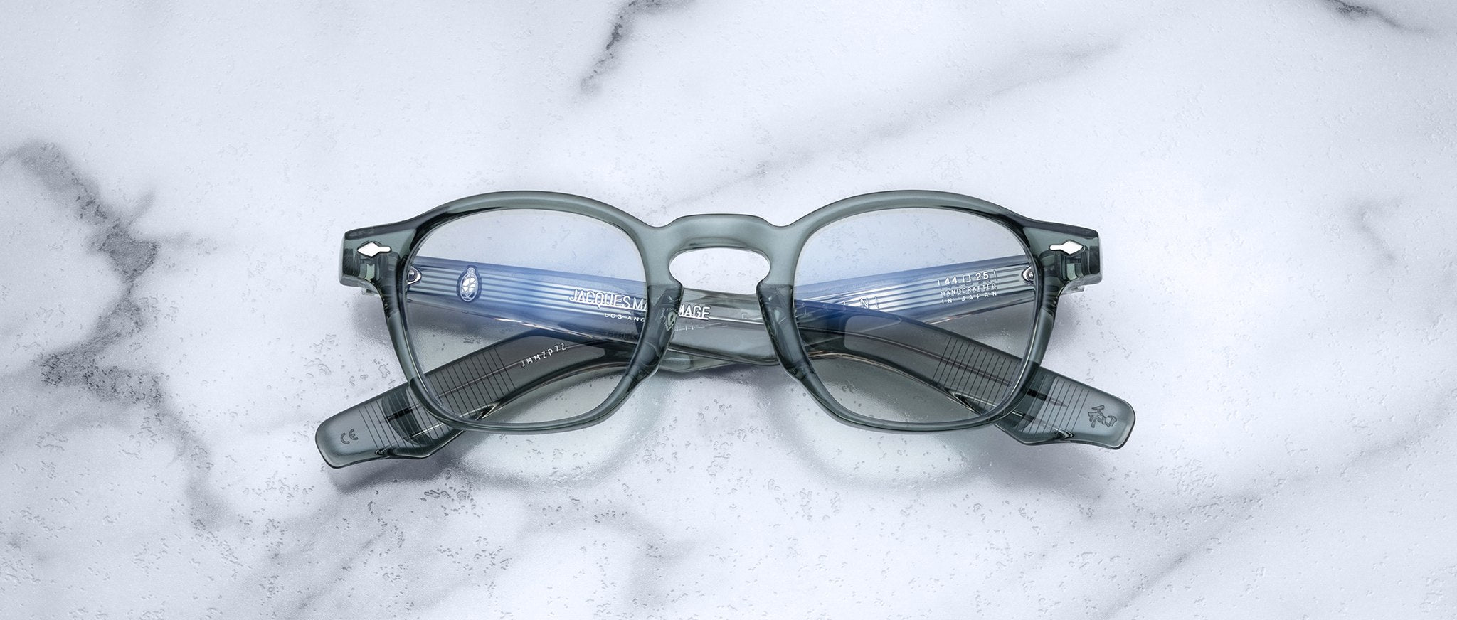 Jacques Marie Mage Zephirin handcrafted translucent gray acetate spectacles with clear lenses rest on white marble, highlighting the brand's refined limited edition eyewear craftsmanship.