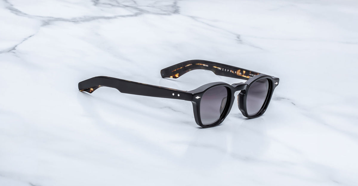 Jacques Marie Mage Zephirin sunglasses in Noir 11, three-quarter view with glossy black acetate, gray-tinted lenses, and subtle tortoiseshell accents on a white marble surface.