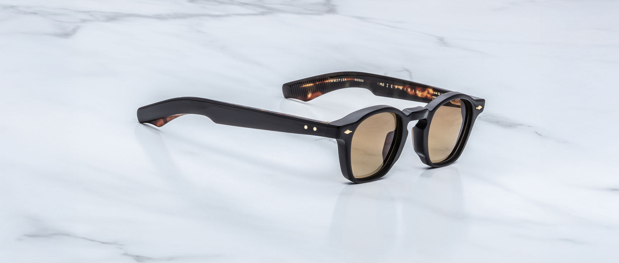 A pair of Jacques Marie Mage Zephirin black sunglasses with brown tinted lenses sits on white marble, highlighting the elegance of limited edition Jacques Marie Mage eyewear.