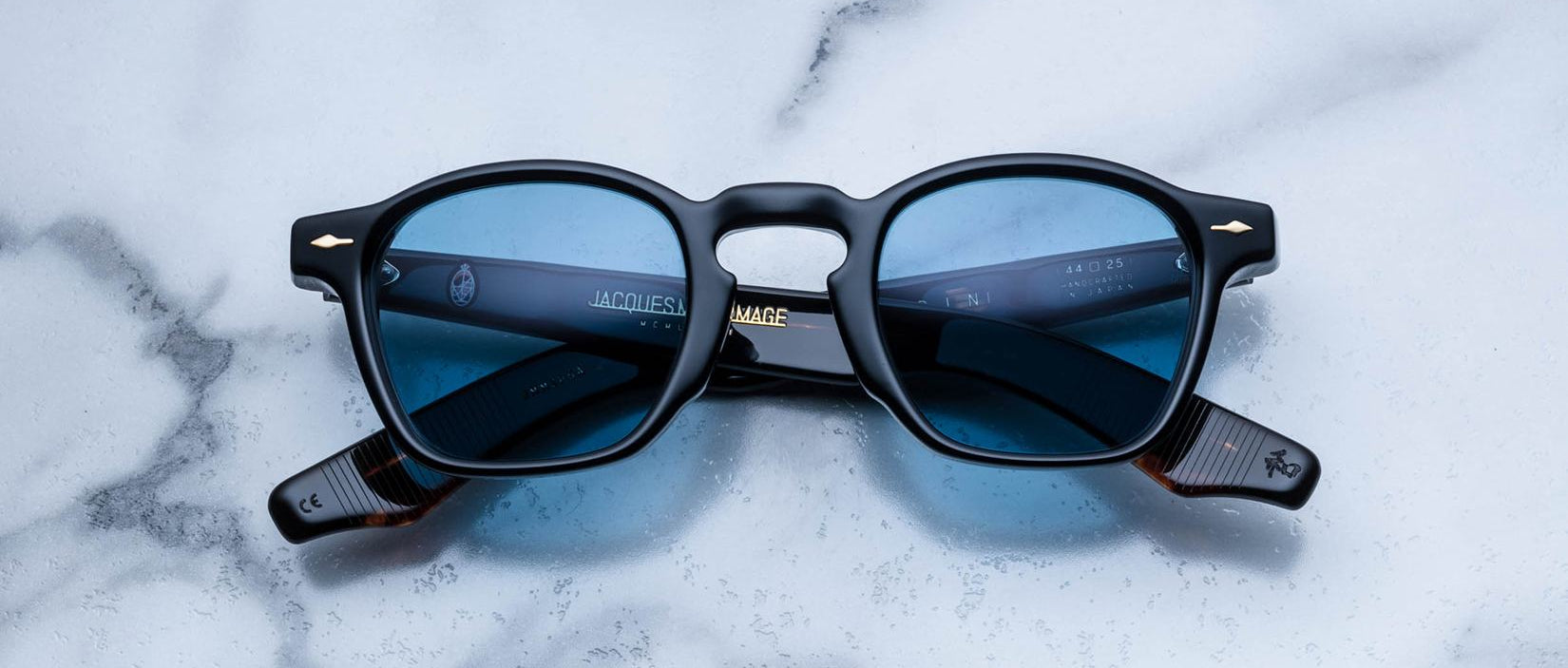 A limited edition pair of Jacques Marie Mage Zephirin black rectangular sunglasses with blue-tinted lenses rests on white marble—luxurious handcrafted acetate eyewear from Jacques Marie Mage.