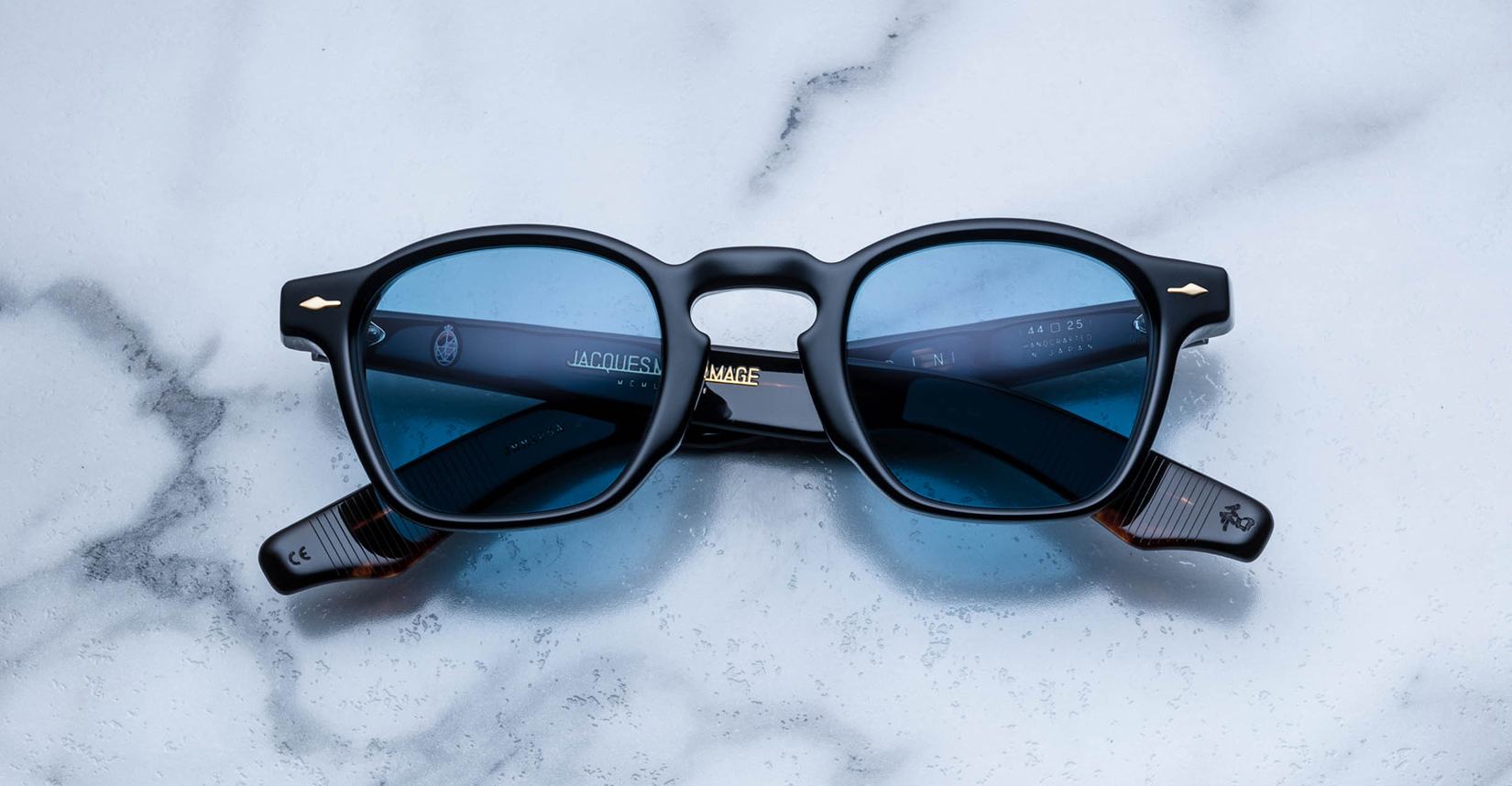A limited edition pair of Jacques Marie Mage Zephirin black rectangular sunglasses with blue-tinted lenses rests on white marble—luxurious handcrafted acetate eyewear from Jacques Marie Mage.