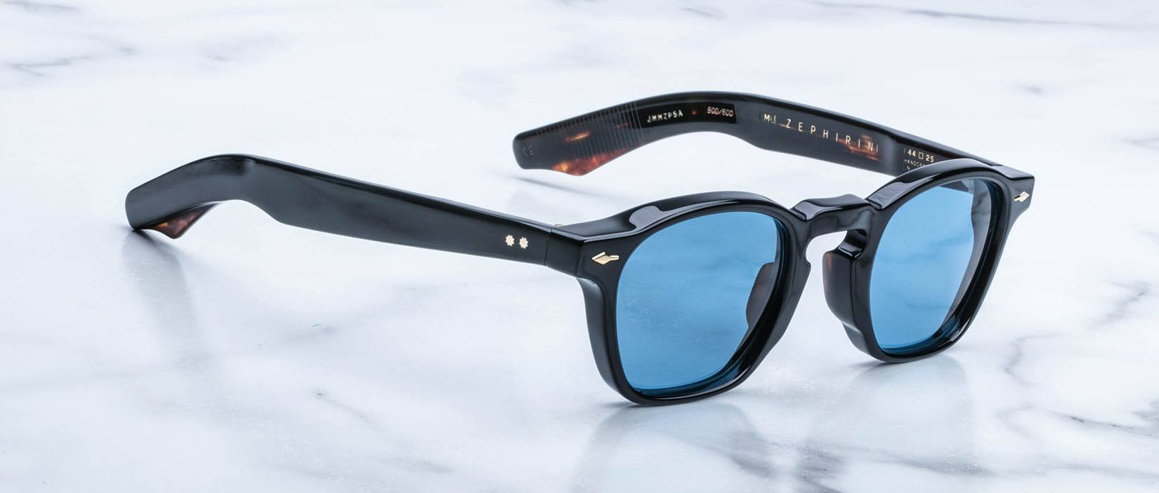 Jacques Marie Mage Zephirin sunglasses feature black acetate, blue lenses, and thick frames, displayed on a white marble surface to highlight the brand’s timeless style in this limited edition eyewear.