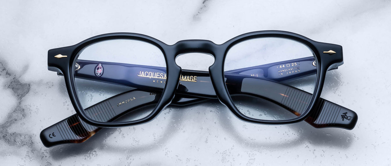 A pair of Jacques Marie Mage Zephirin black rectangular eyeglasses with gold accents, featuring handcrafted acetate frames, are folded and displayed on a white marble surface.