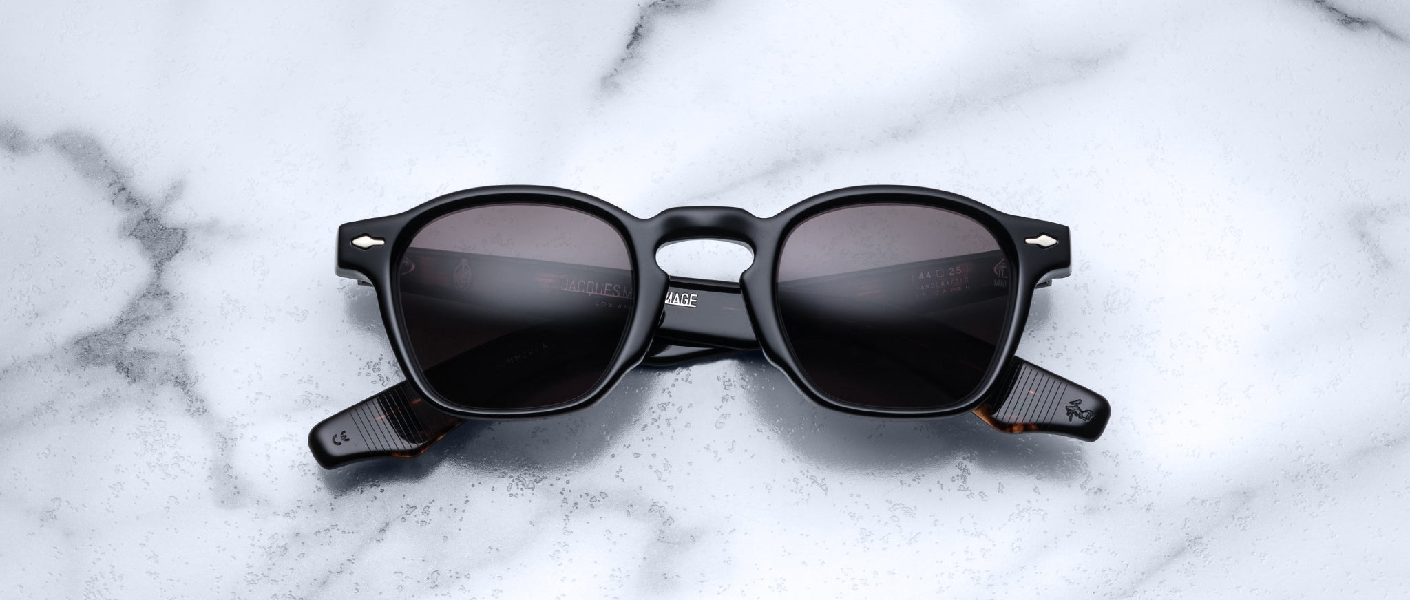 A pair of Jacques Marie Mage Zephirin sunglasses with black square frames and dark lenses sits on white marble, highlighting the brand’s limited edition craftsmanship.
