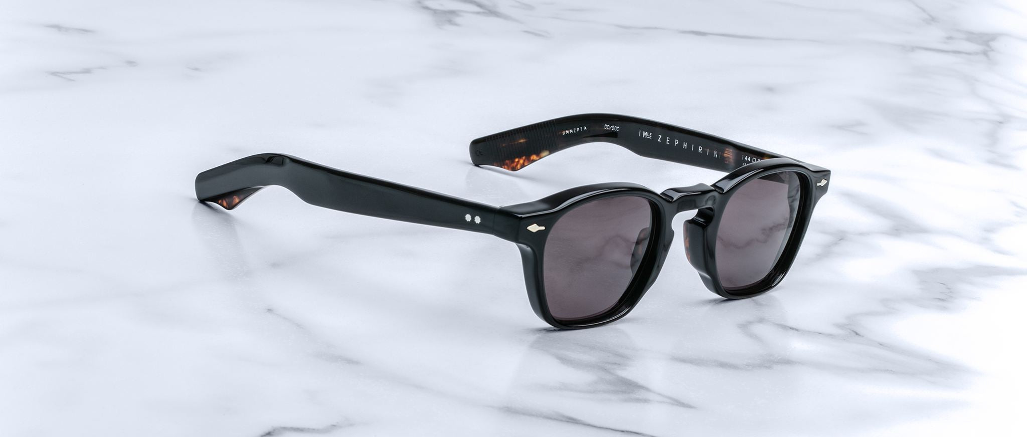 Jacques Marie Mage Zephirin sunglasses feature a classic handcrafted acetate frame with dark lenses, inspired by the brand’s limited edition eyewear, and are displayed on a white marble surface.