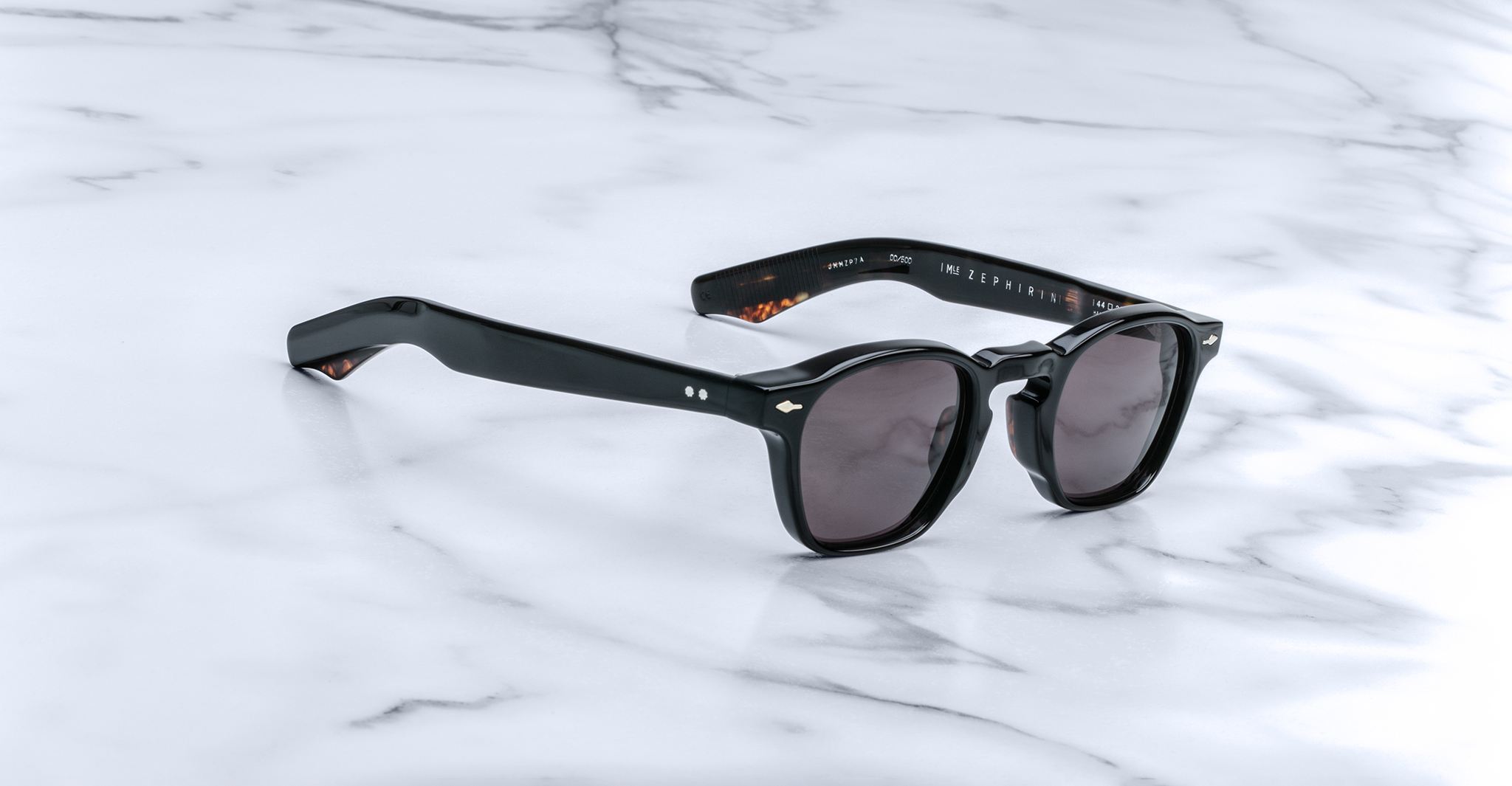 Jacques Marie Mage Zephirin sunglasses feature a classic handcrafted acetate frame with dark lenses, inspired by the brand’s limited edition eyewear, and are displayed on a white marble surface.