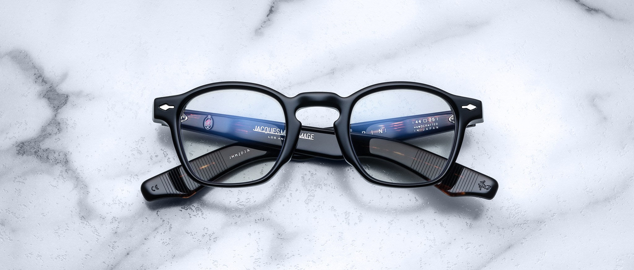 A pair of Jacques Marie Mage Zephirin eyeglasses by Jacques Marie Mage, featuring handcrafted black acetate and round lenses, rests on a white marble surface with reflected light in the lenses.