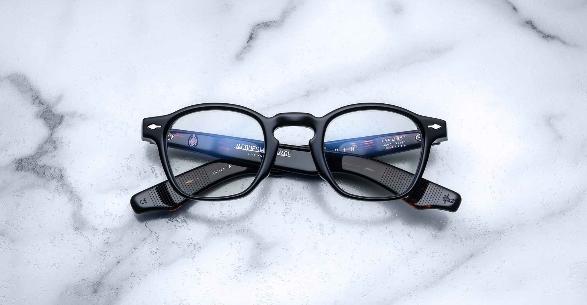 A pair of Jacques Marie Mage Zephirin eyeglasses by Jacques Marie Mage, featuring handcrafted black acetate and round lenses, rests on a white marble surface with reflected light in the lenses.