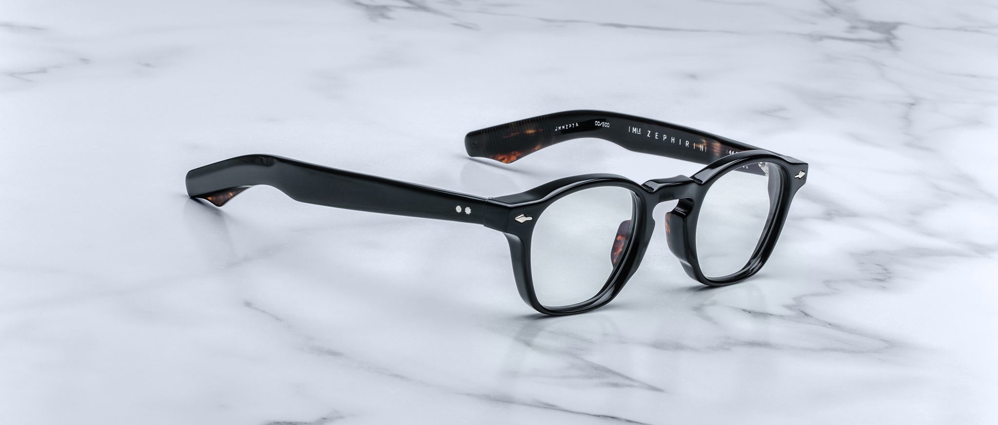 A pair of Jacques Marie Mage Zephirin eyeglasses with black thick rims and clear lenses rests on a white marble surface.