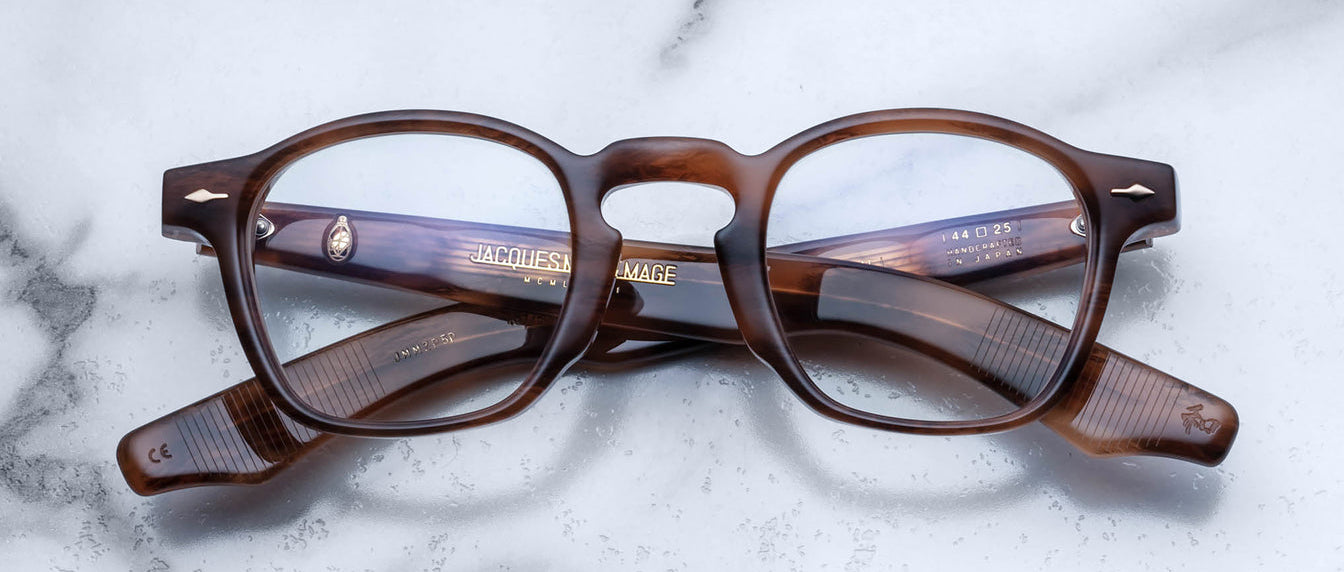 A pair of Jacques Marie Mage Zephirin handcrafted acetate spectacles in brown tortoiseshell with clear lenses, folded and resting on a white marble surface.