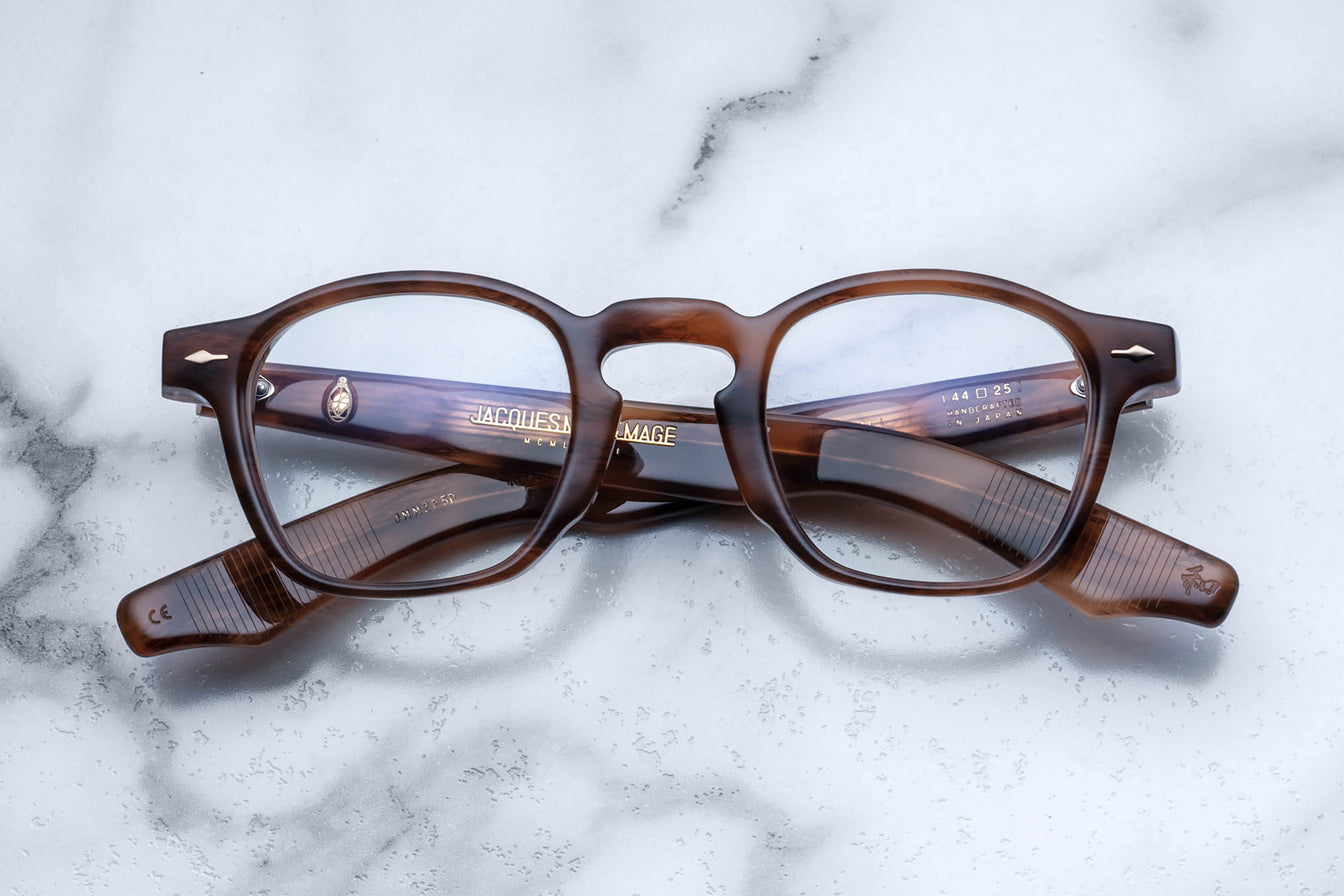 A pair of Jacques Marie Mage Zephirin handcrafted acetate spectacles in brown tortoiseshell with clear lenses, folded and resting on a white marble surface.