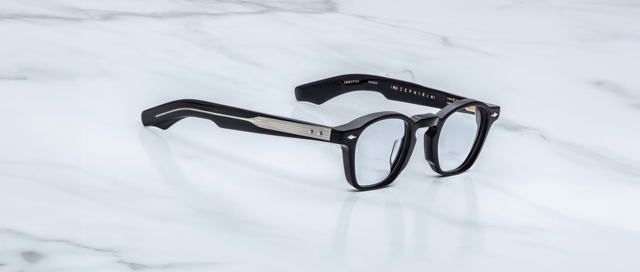 Jacques Marie Mage Zephirin eyeglasses by Jacques Marie Mage, featuring black frames and metal temple detailing, are displayed on white marble, highlighting the handcrafted acetate and striking limited edition design.