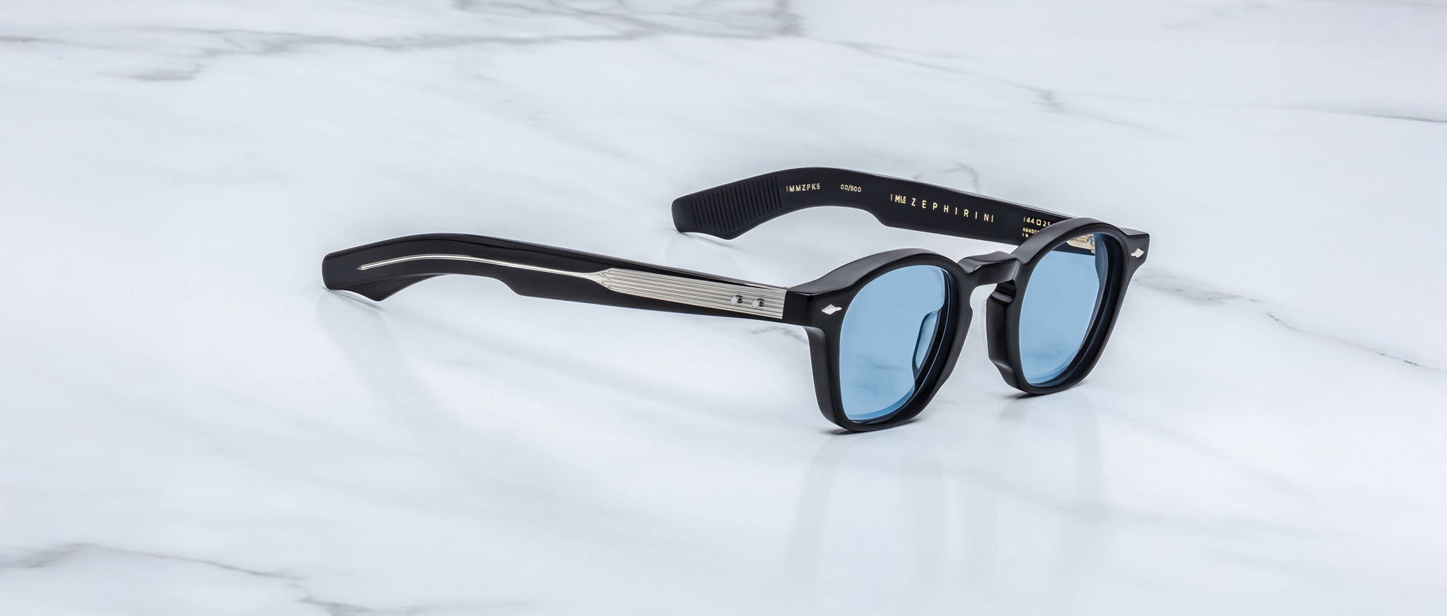 A pair of Jacques Marie Mage Zephirin sunglasses with black frames and blue-tinted lenses is displayed on a white marble surface, highlighting the exclusive style of this limited edition Jacques Marie Mage eyewear.
