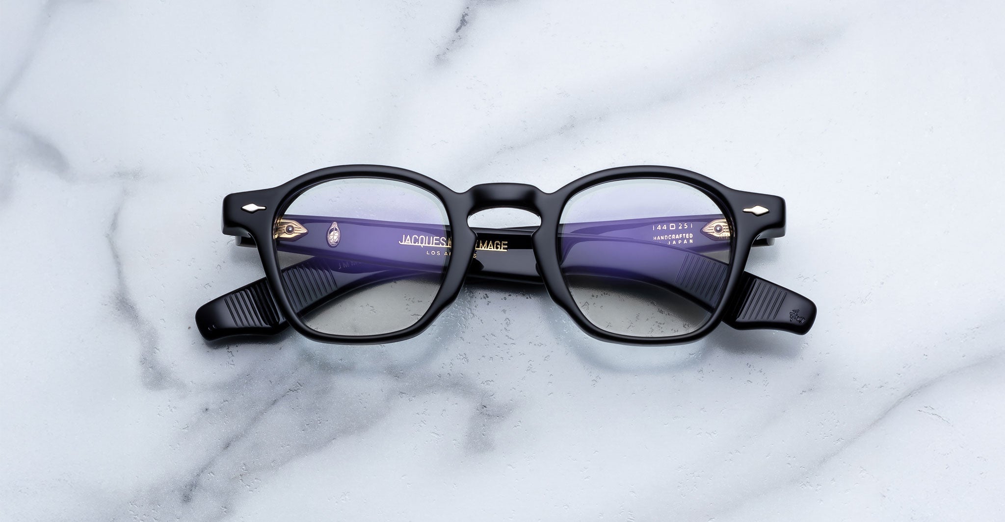 A folded pair of Jacques Marie Mage Zephirin black-rimmed eyeglasses with round lenses rests on a white marble surface—handcrafted acetate frames by Jacques Marie Mage that exude timeless style.