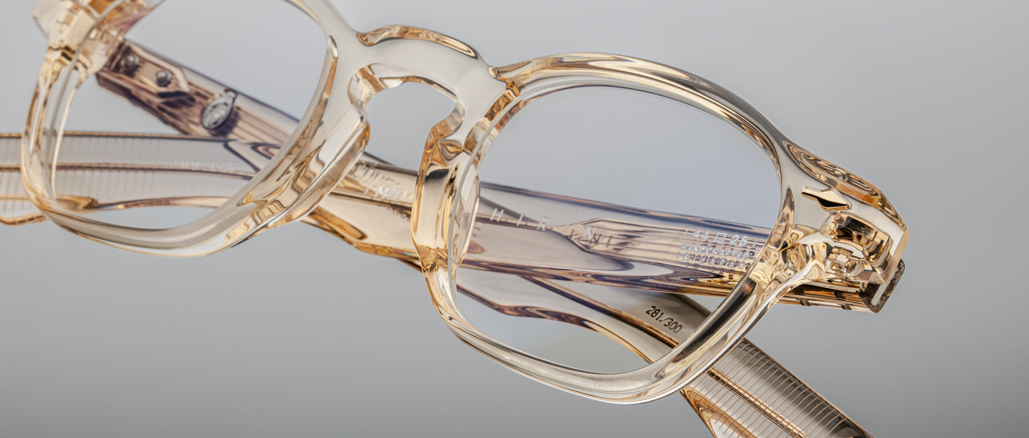 A close-up of Jacques Marie Mage Zephirin spectacles—transparent, beige-tinted handcrafted acetate frames with clear lenses and patterned arms—rests on a reflective surface against a plain background.