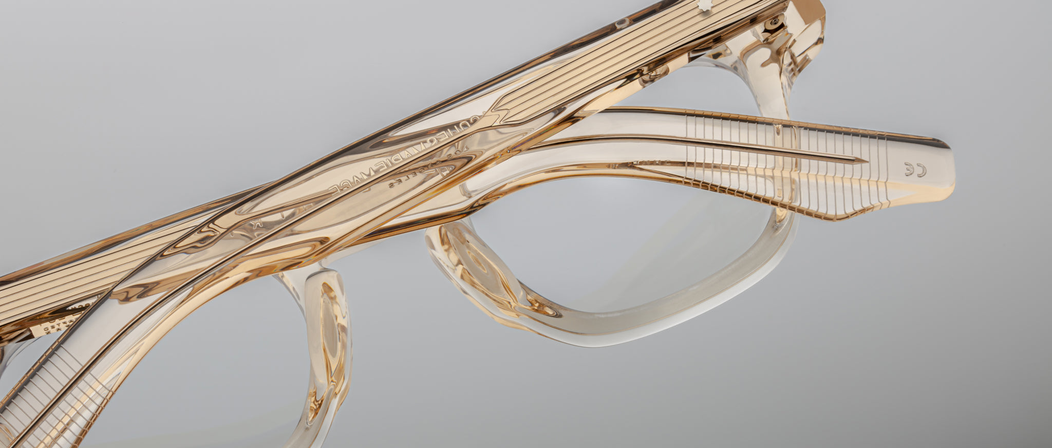 Close-up of Jacques Marie Mage Zephirin eyeglass frames, featuring clear acetate, gold accents, and fine etched details by Jacques Marie Mage. These limited edition pieces are showcased against a plain gray background.