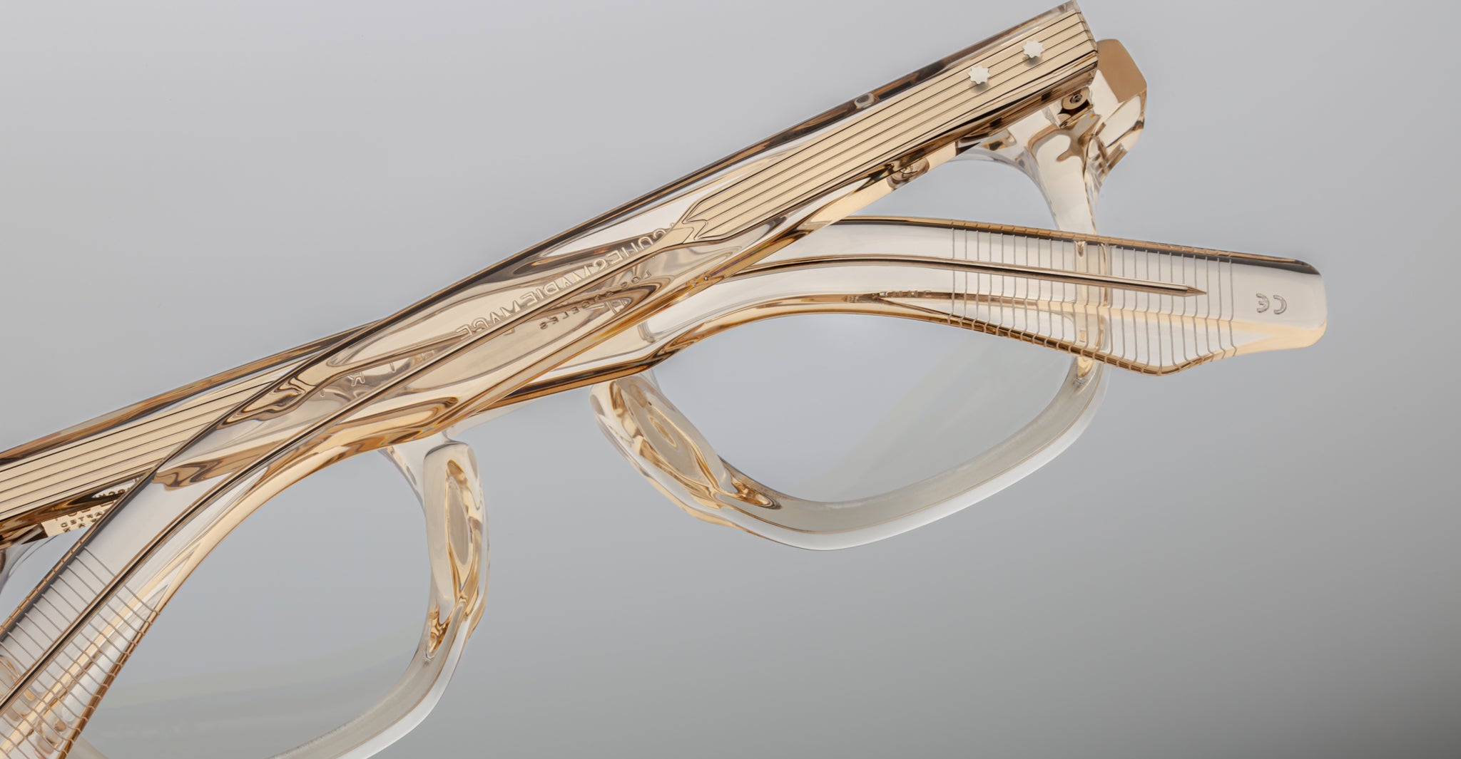 Close-up of Jacques Marie Mage Zephirin eyeglass frames, featuring clear acetate, gold accents, and fine etched details by Jacques Marie Mage. These limited edition pieces are showcased against a plain gray background.