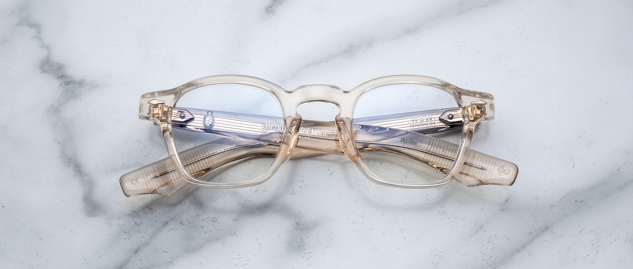Jacques Marie Mage Zephirin eyeglasses by Jacques Marie Mage, featuring round lenses and transparent frames, are folded and placed on a white marble surface.