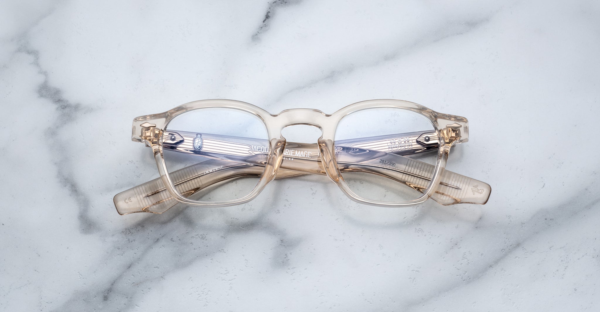 Jacques Marie Mage Zephirin eyeglasses by Jacques Marie Mage, featuring round lenses and transparent frames, are folded and placed on a white marble surface.