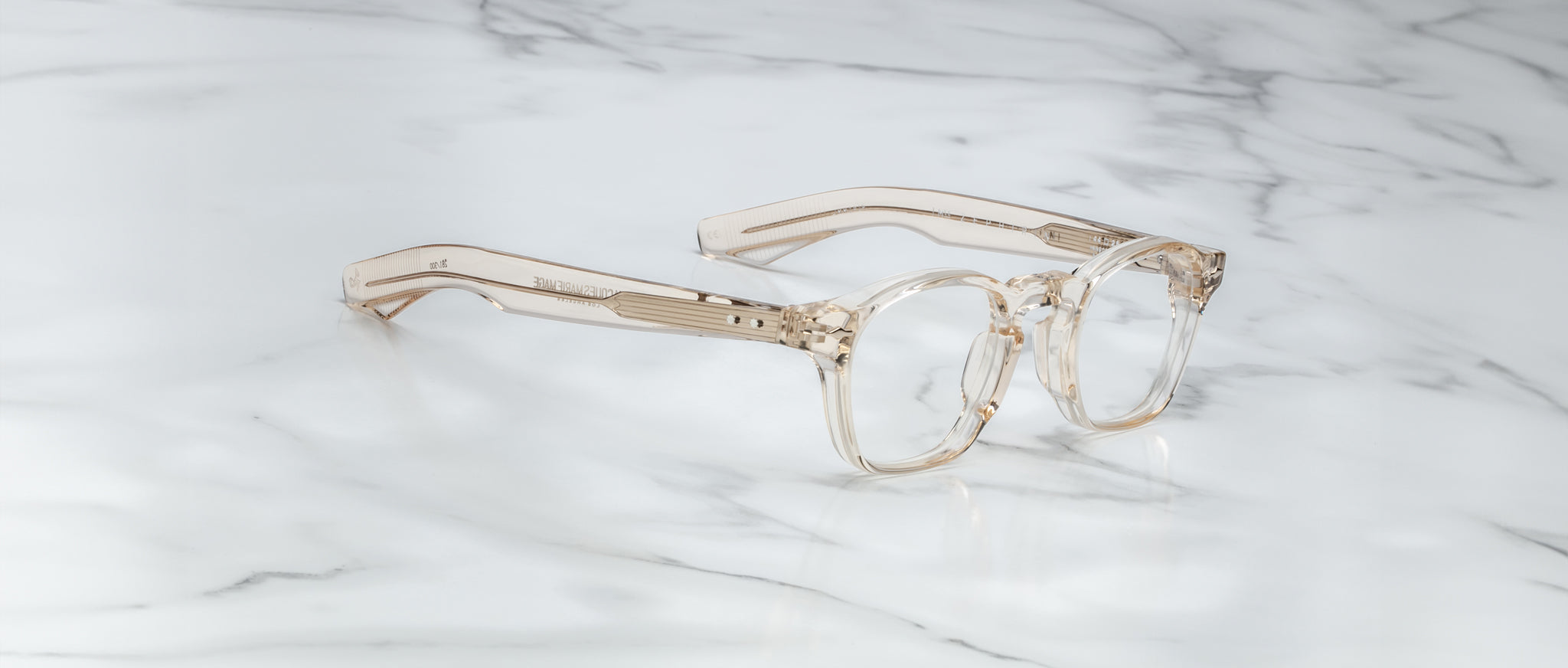 A pair of limited edition Jacques Marie Mage Zephirin eyeglasses in clear, beige-tinted handcrafted acetate with thick frames rests on white marble, showcasing the brand’s signature design and luxury craftsmanship.