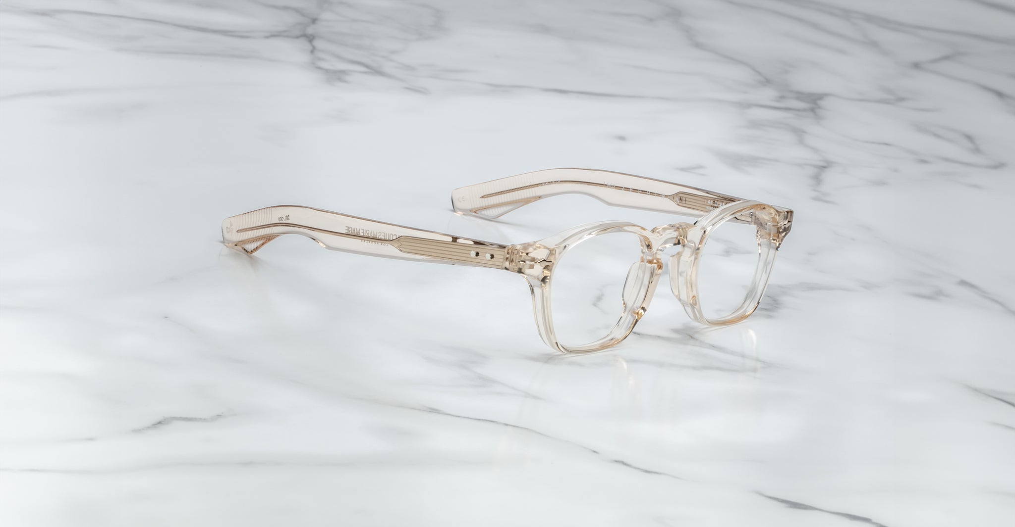 A pair of limited edition Jacques Marie Mage Zephirin eyeglasses in clear, beige-tinted handcrafted acetate with thick frames rests on white marble, showcasing the brand’s signature design and luxury craftsmanship.