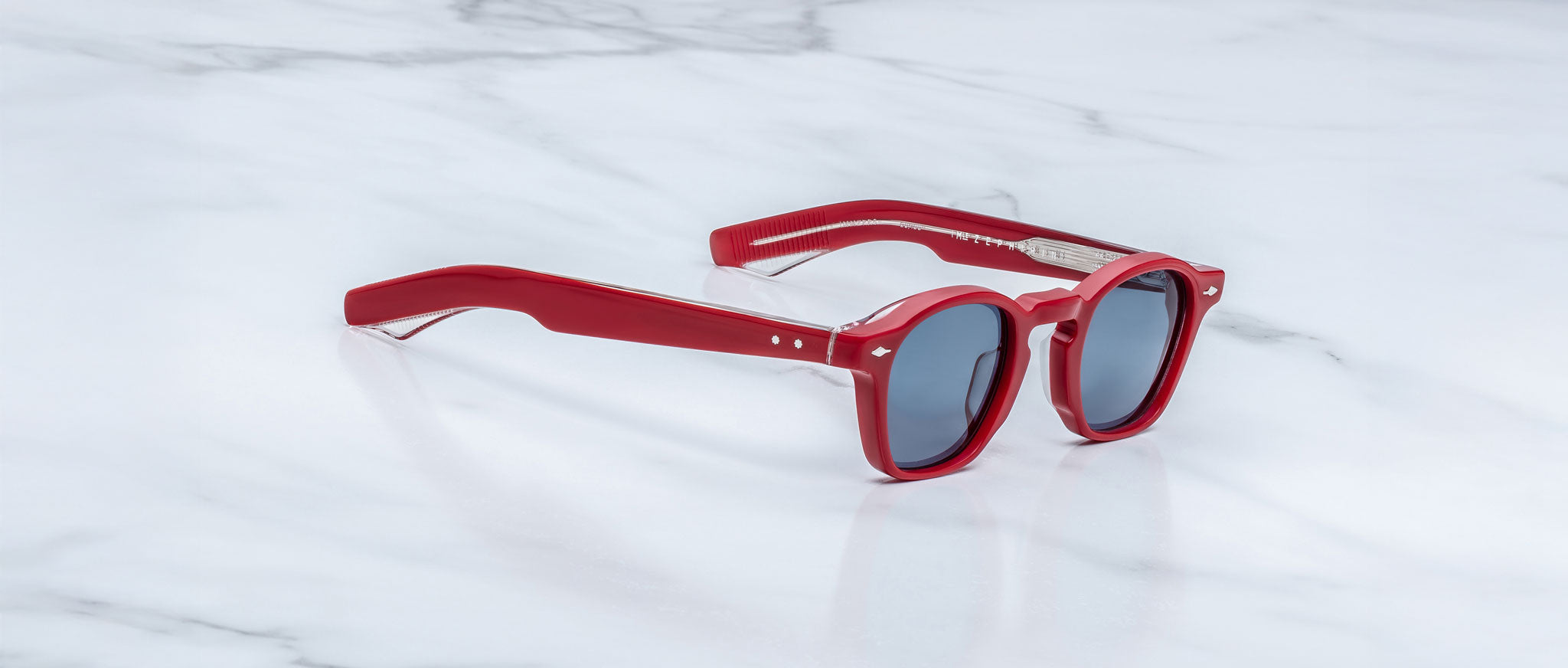 Limited edition Jacques Marie Mage Zephirin sunglasses with red frames and dark lenses rest stylishly on a white marble surface.