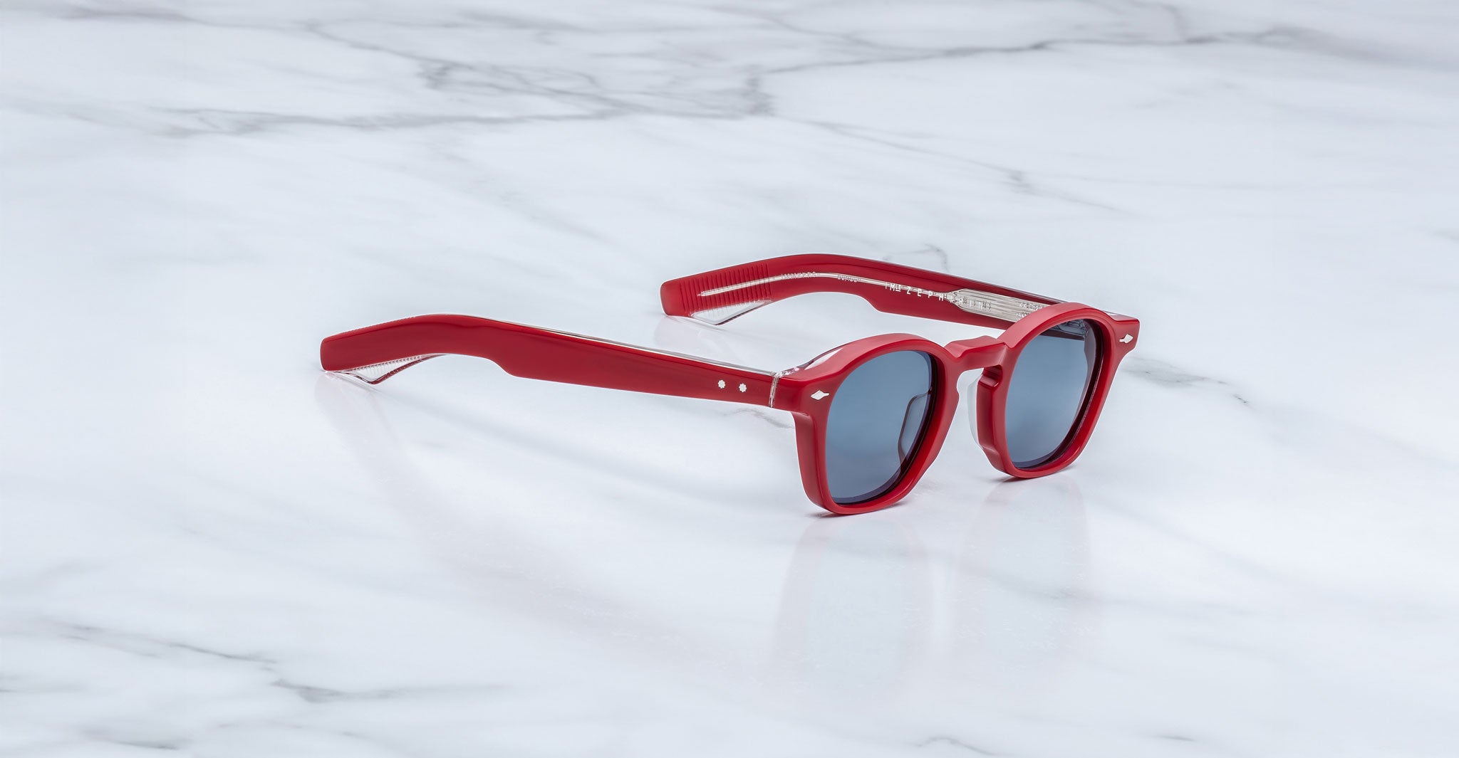 Limited edition Jacques Marie Mage Zephirin sunglasses with red frames and dark lenses rest stylishly on a white marble surface.