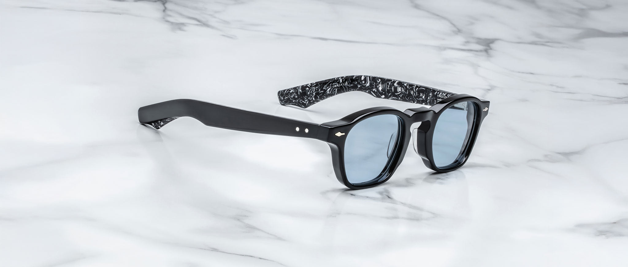 Jacques Marie Mage Zephirin sunglasses feature blue-tinted lenses and marbled inner temples in handcrafted acetate, beautifully displayed on white marble—a striking limited edition by Jacques Marie Mage.
