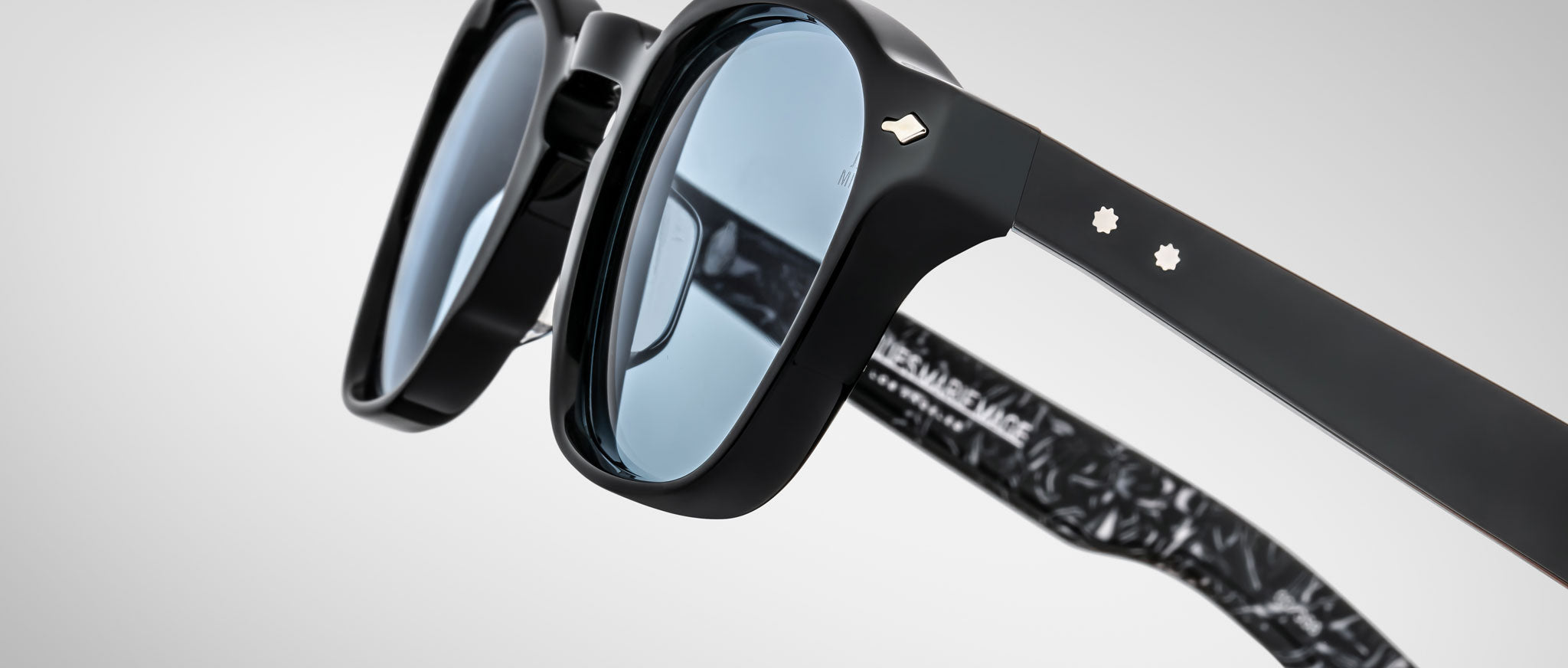 Close-up of Jacques Marie Mage Zephirin sunglasses—limited edition handcrafted acetate eyewear in black with blue tinted lenses, metal rivets on the arms, and a marbled black inner temple pattern.