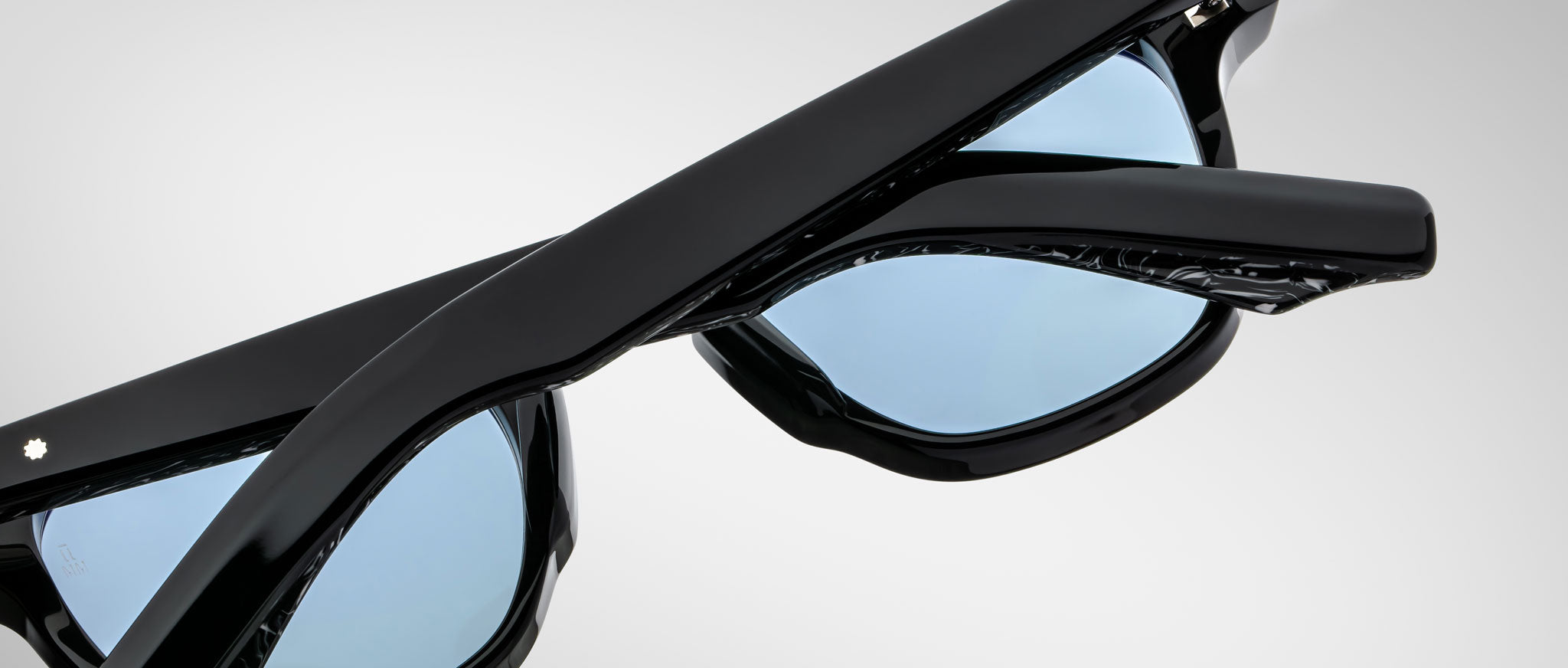 Folded Jacques Marie Mage Zephirin black sunglasses with blue-tinted lenses, inspired by the limited edition eyewear collection, displayed on a plain light grey background.