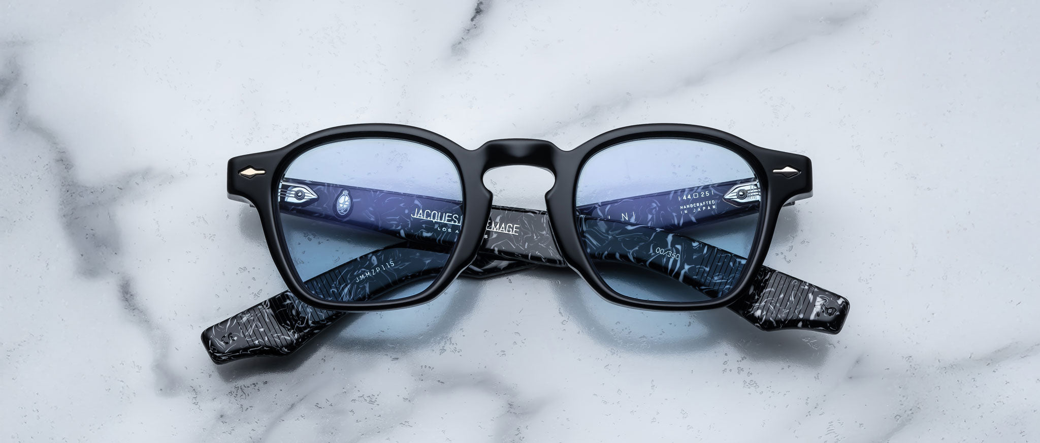 A pair of Jacques Marie Mage Zephirin black-framed eyeglasses with blue-tinted lenses rests on white marble, highlighting the refined craftsmanship of Jacques Marie Mage.