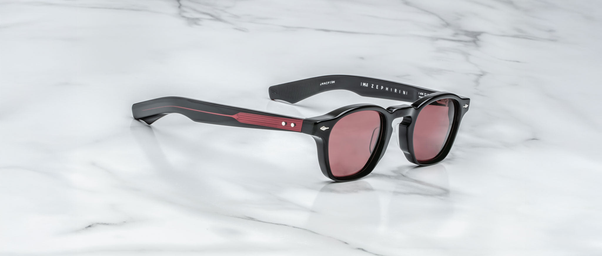 A pair of Jacques Marie Mage Zephirin limited edition black sunglasses with red-tinted lenses and red accents on the arms, inspired by Jacques Marie Mage, displayed on a white marble surface.