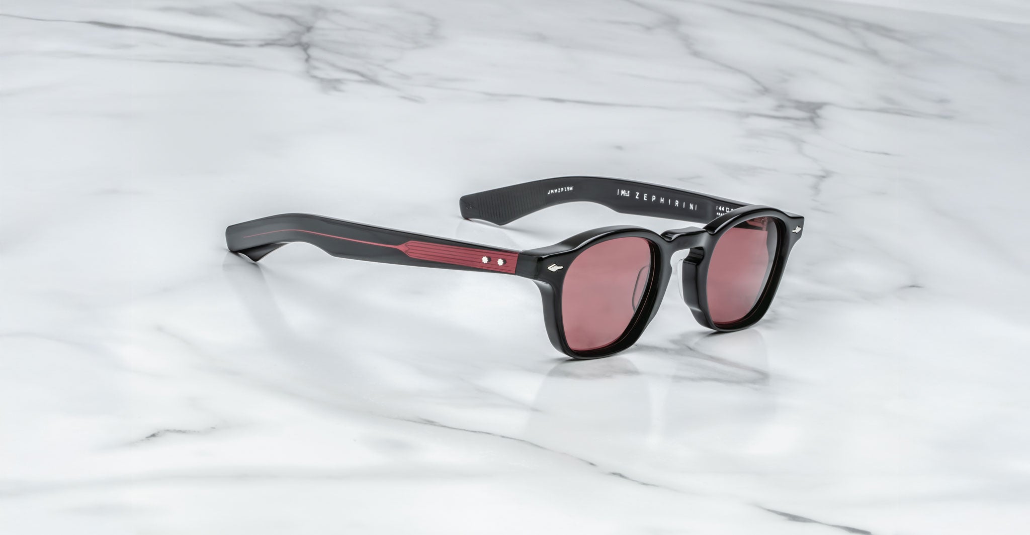 A pair of Jacques Marie Mage Zephirin limited edition black sunglasses with red-tinted lenses and red accents on the arms, inspired by Jacques Marie Mage, displayed on a white marble surface.