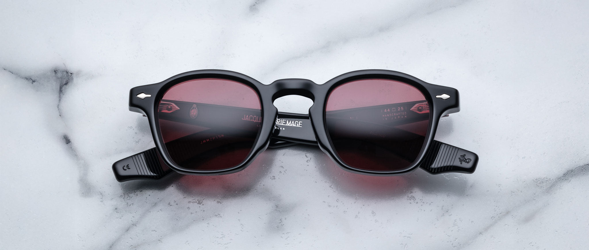 Jacques Marie Mage Zephirin sunglasses in black with dark red lenses are displayed open on white marble—a stylish presentation of this limited edition Jacques Marie Mage eyewear.