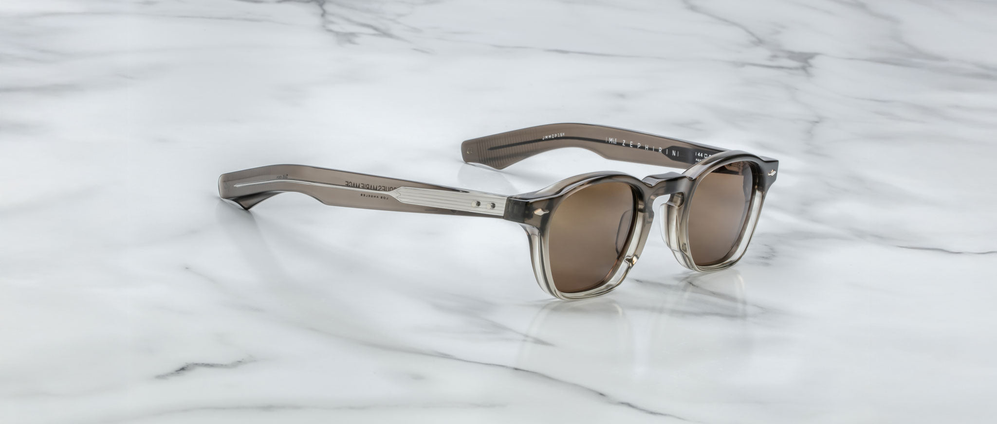A pair of Jacques Marie Mage Zephirin sunglasses with brown-tinted lenses and transparent gray frames rests on white marble, highlighting this limited edition Jacques Marie Mage eyewear.