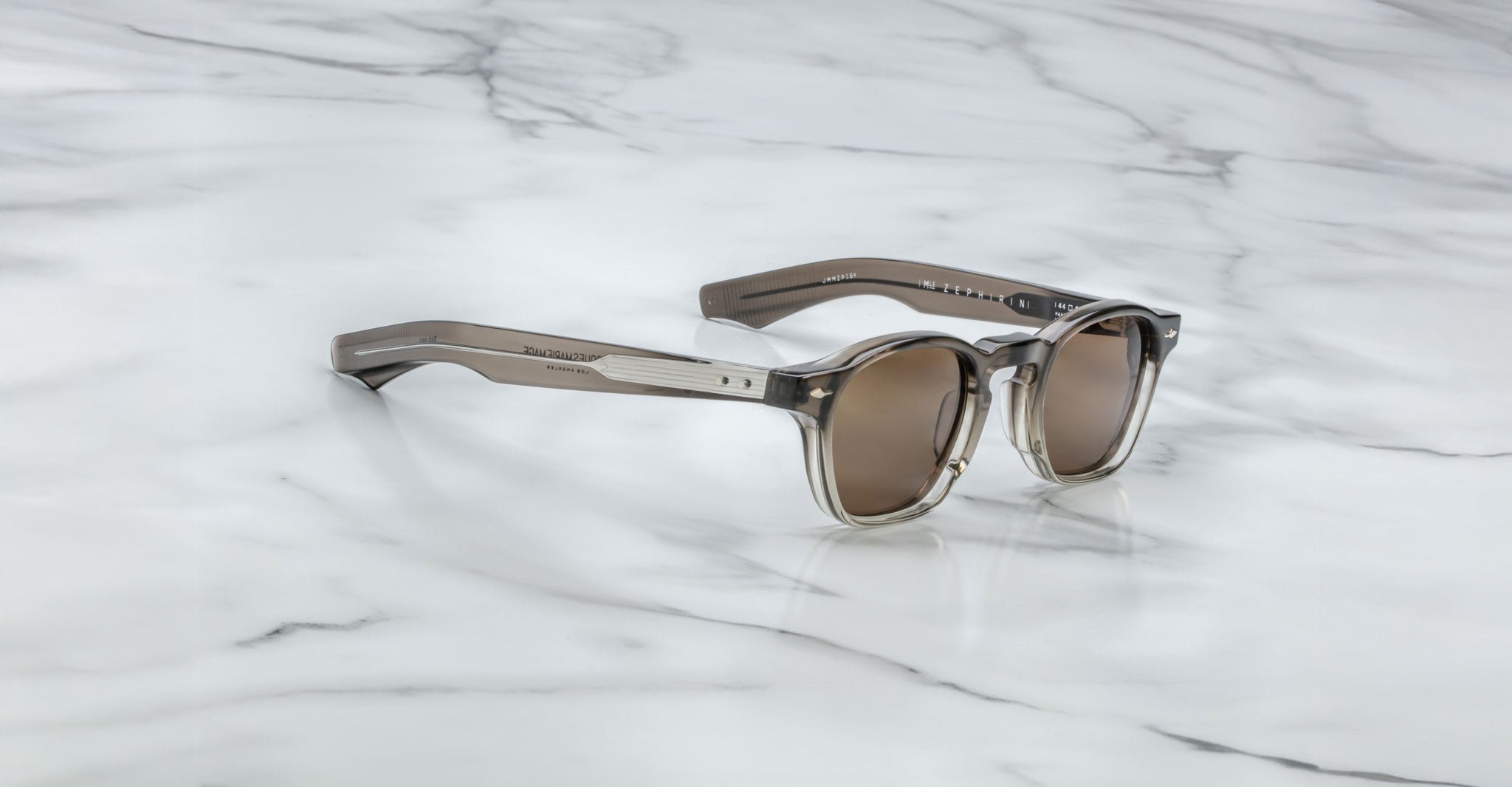 A pair of Jacques Marie Mage Zephirin sunglasses with brown-tinted lenses and transparent gray frames rests on white marble, highlighting this limited edition Jacques Marie Mage eyewear.