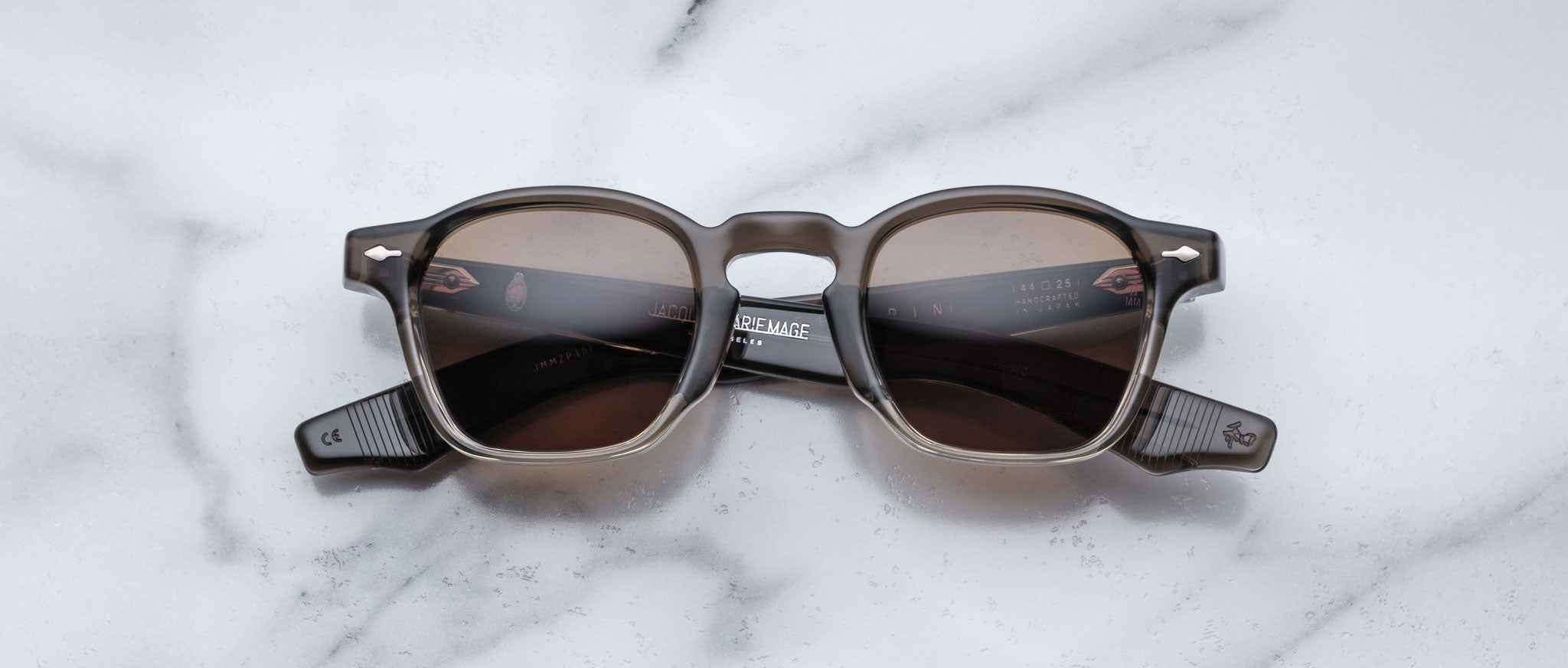 A pair of square, brown-tinted Jacques Marie Mage Zephirin sunglasses with thick dark frames rests on white marble—a luxurious limited edition from Jacques Marie Mage for discerning collectors.