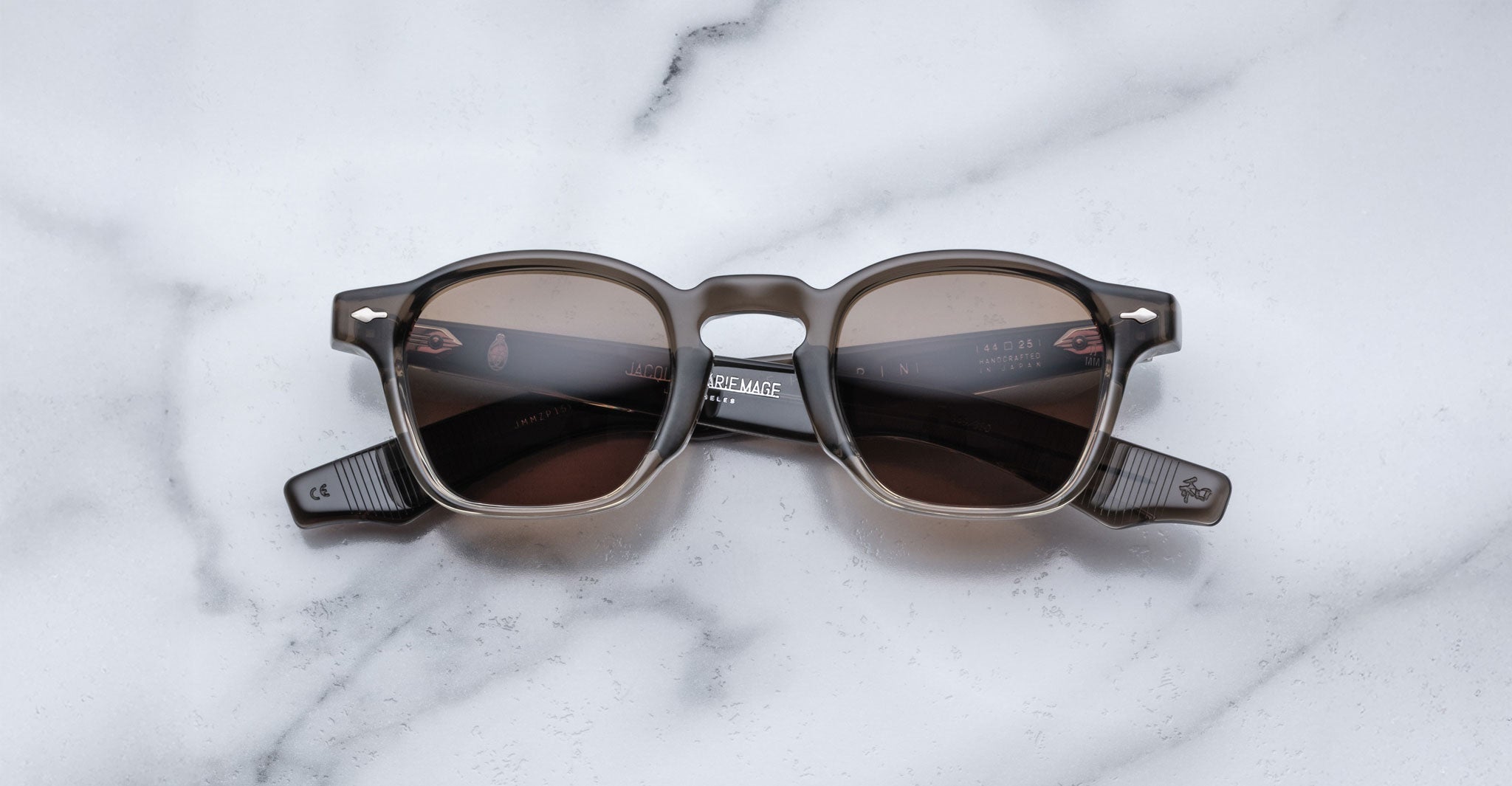 A pair of square, brown-tinted Jacques Marie Mage Zephirin sunglasses with thick dark frames rests on white marble—a luxurious limited edition from Jacques Marie Mage for discerning collectors.