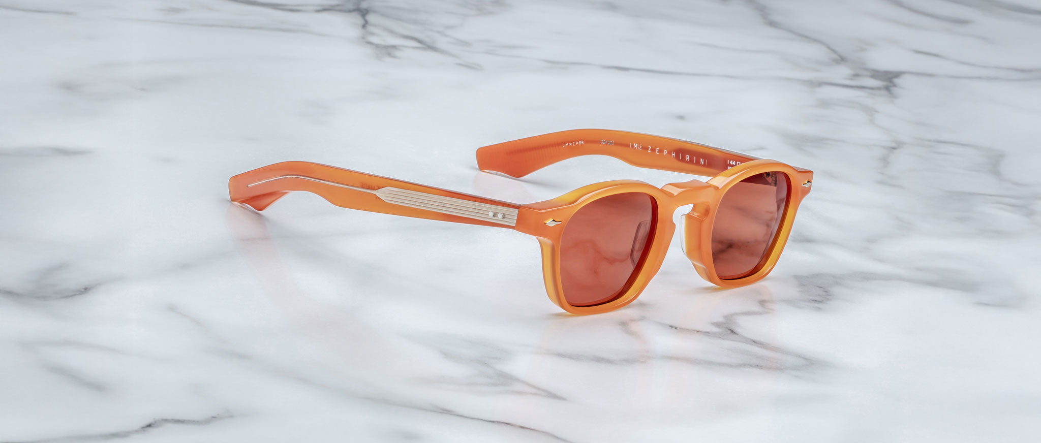 The Jacques Marie Mage Zephirin sunglasses, featuring orange frames and red-tinted lenses, are displayed on a white marble surface, highlighting the brand’s signature handcrafted acetate style.