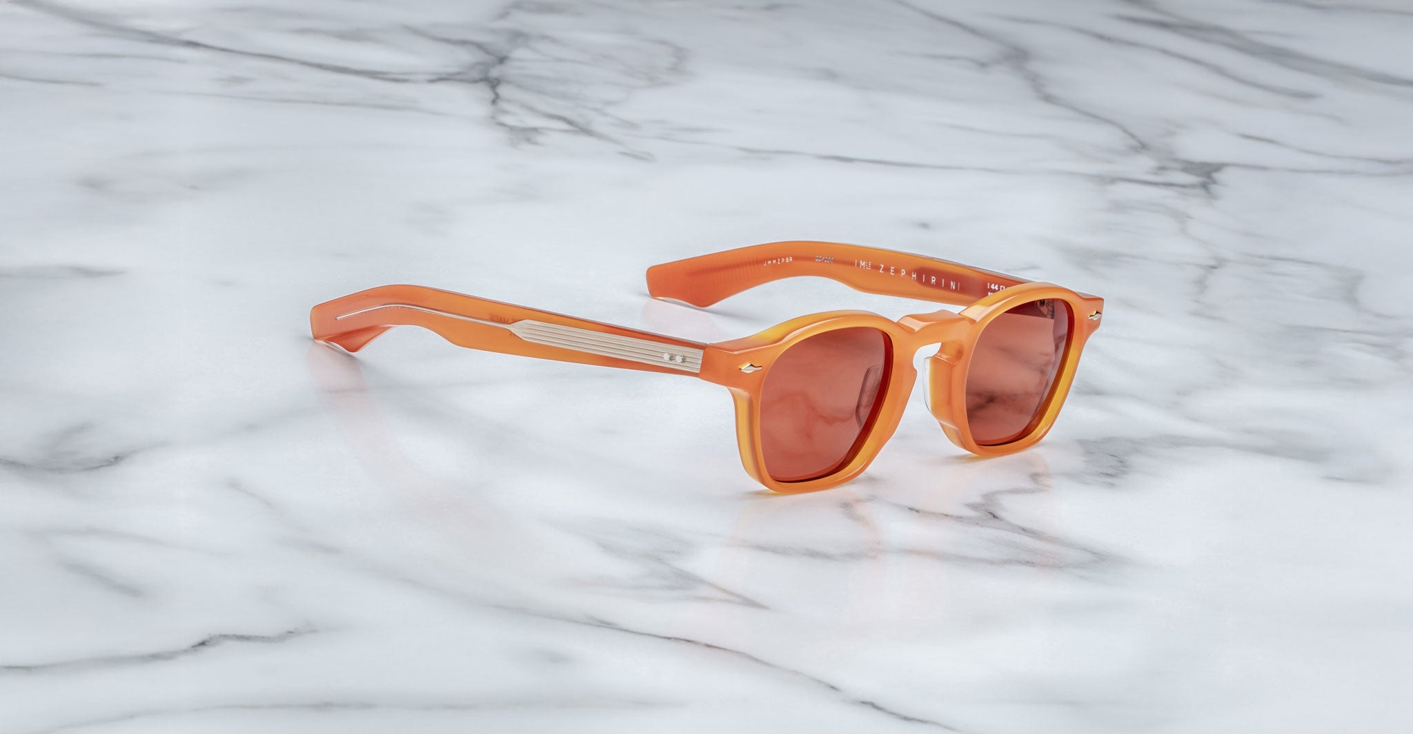 The Jacques Marie Mage Zephirin sunglasses, featuring orange frames and red-tinted lenses, are displayed on a white marble surface, highlighting the brand’s signature handcrafted acetate style.