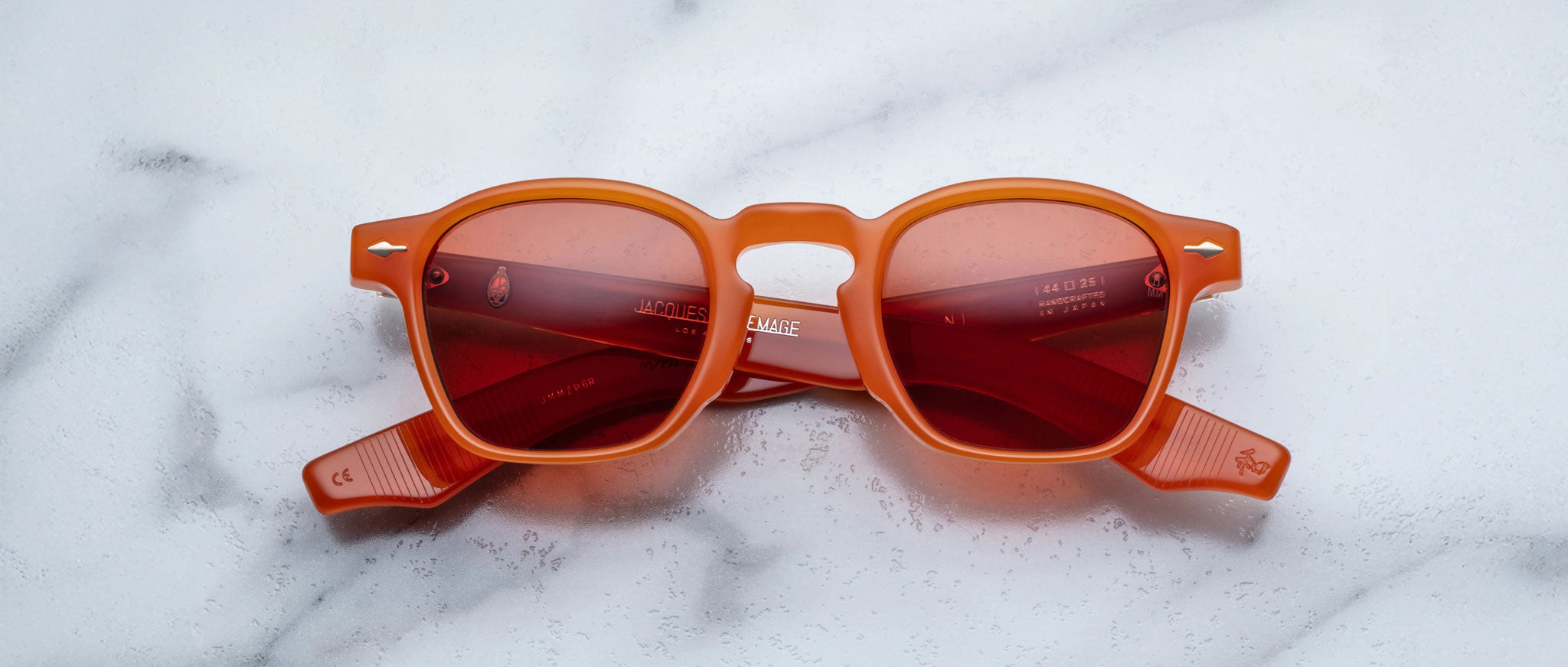 Jacques Marie Mage Zephirin limited edition eyewear, featuring orange frames and red-tinted lenses, is displayed on white marble, showcasing the bold craftsmanship of Jacques Marie Mage.