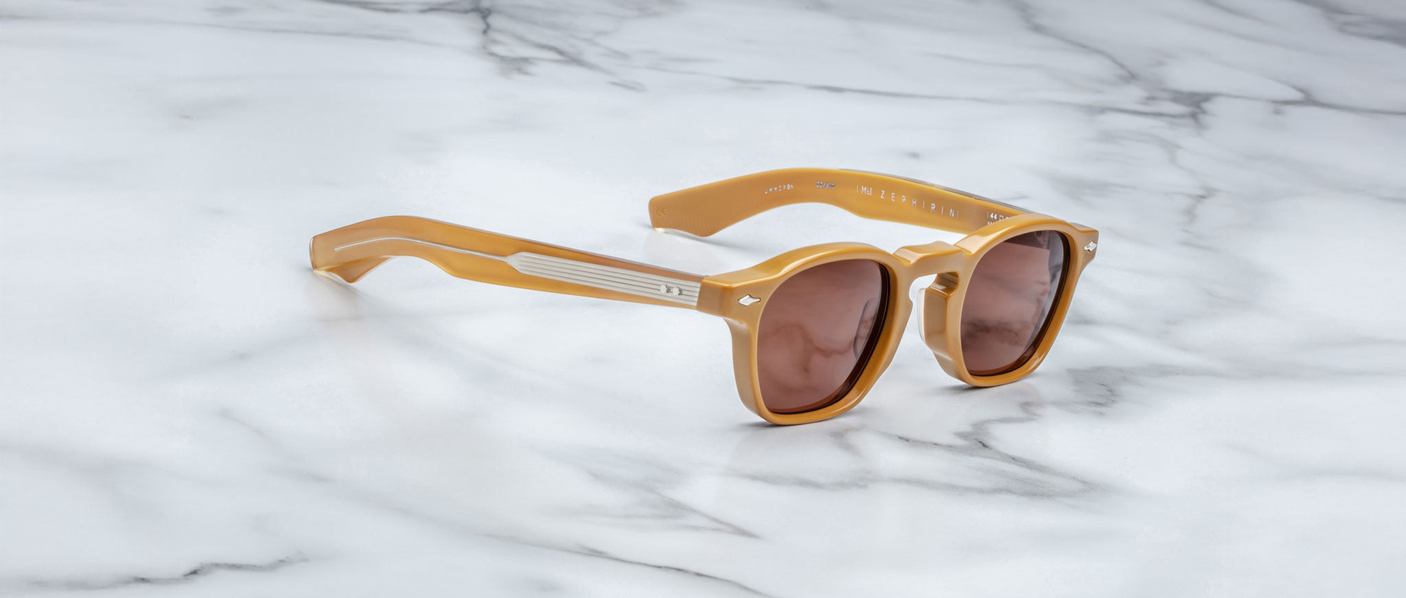 A pair of Jacques Marie Mage Zephirin sunglasses with tan frames and brown lenses rests on a white marble surface.